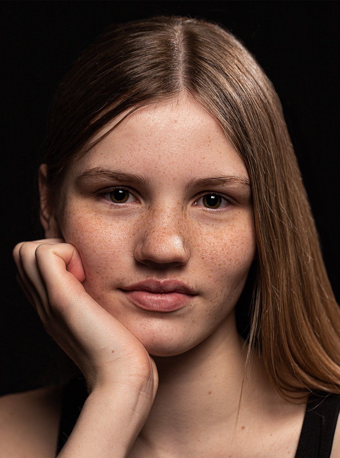 Studio portrait of a teenage girl with freckles and straight brown hair posing against a black background.