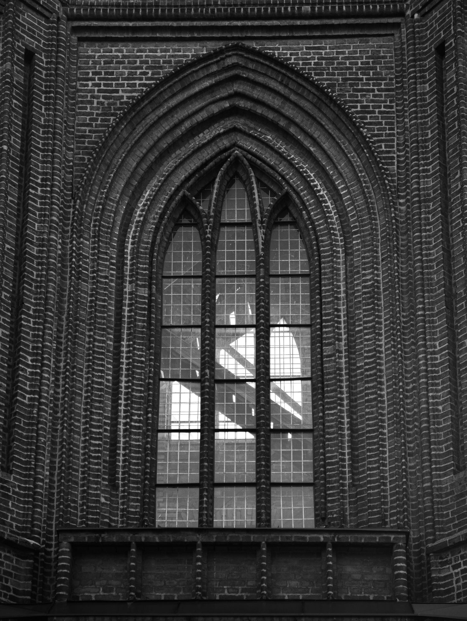 Black and white image of a tall Gothic arched window set in a detailed brick church wall.