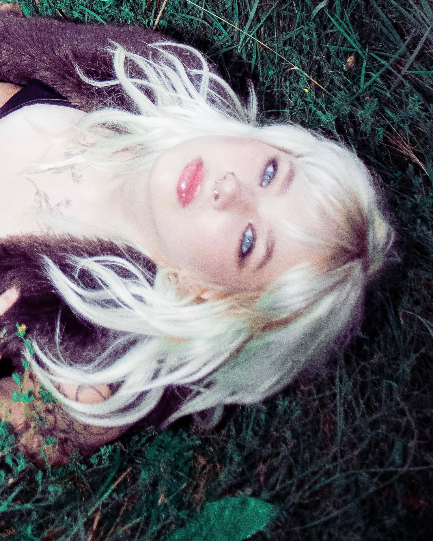 Platinum blonde woman with blue eyes and tattoos lying in grass, gazing upward in a dreamy outdoor portrait.