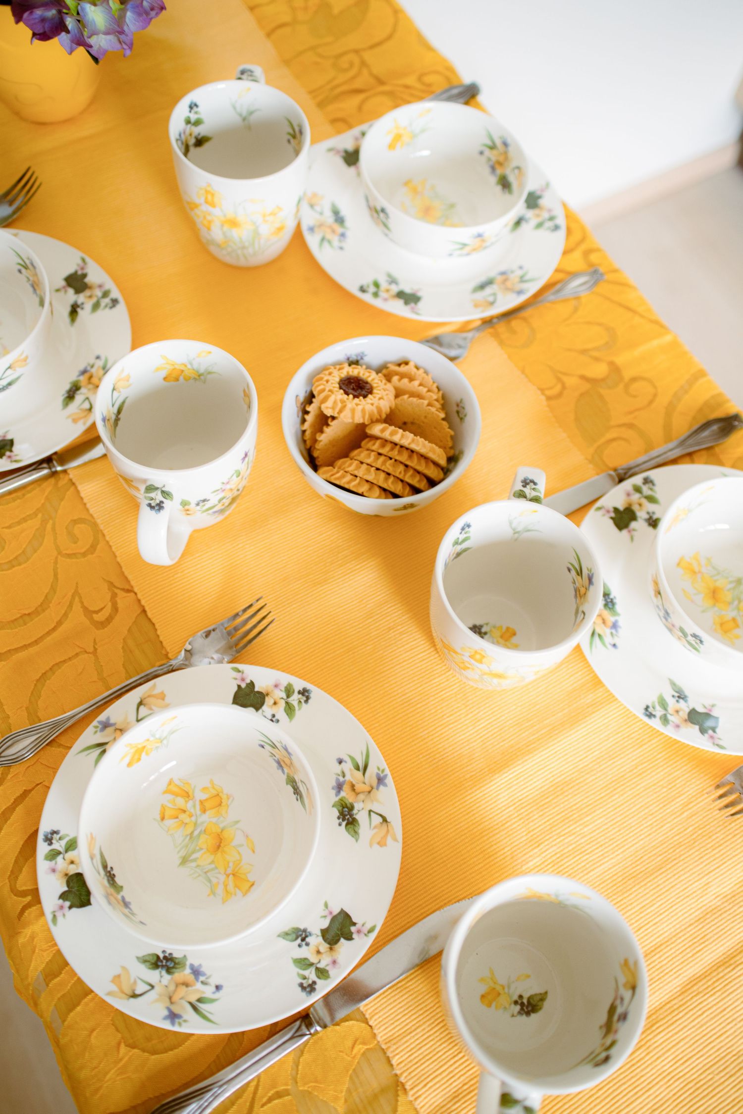 Yellow floral tea set with white porcelain cups, plates, and a bowl of cookies on a yellow tablecloth.