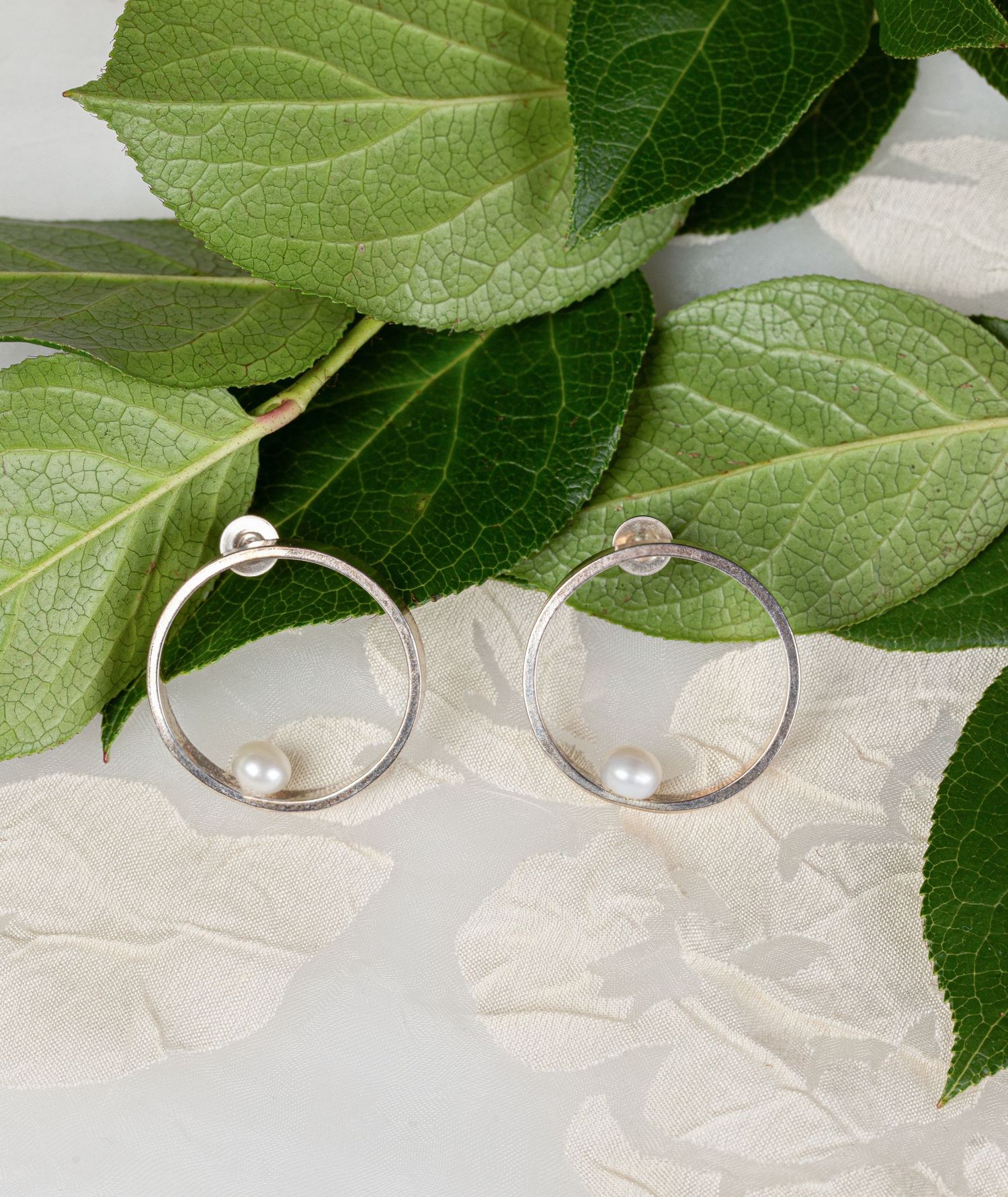 Silver hoop earrings with white freshwater pearls displayed on green leaves and white petals.