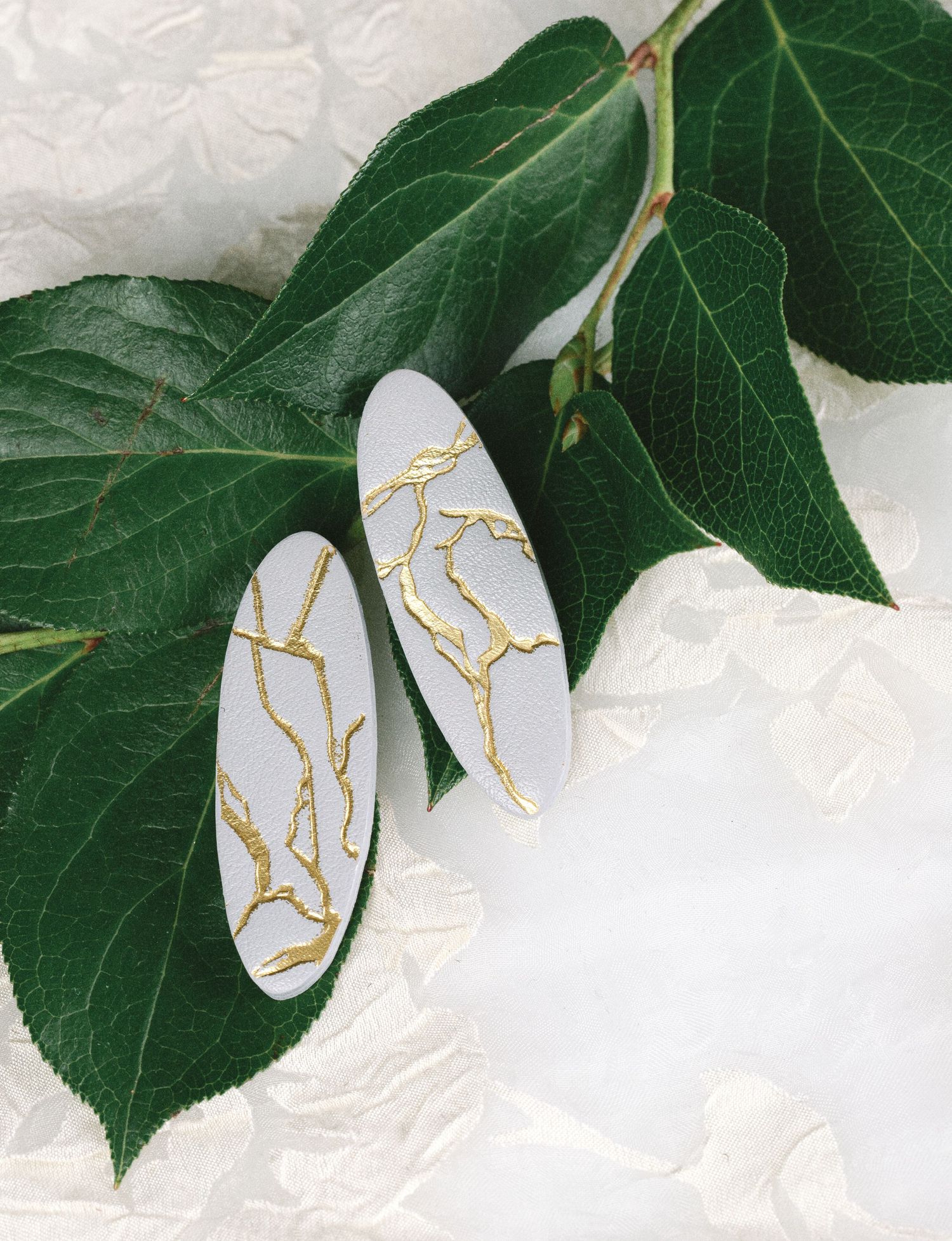 White oval earrings with gold kintsugi-inspired crack details, displayed on lush green ivy leaves against white lace.