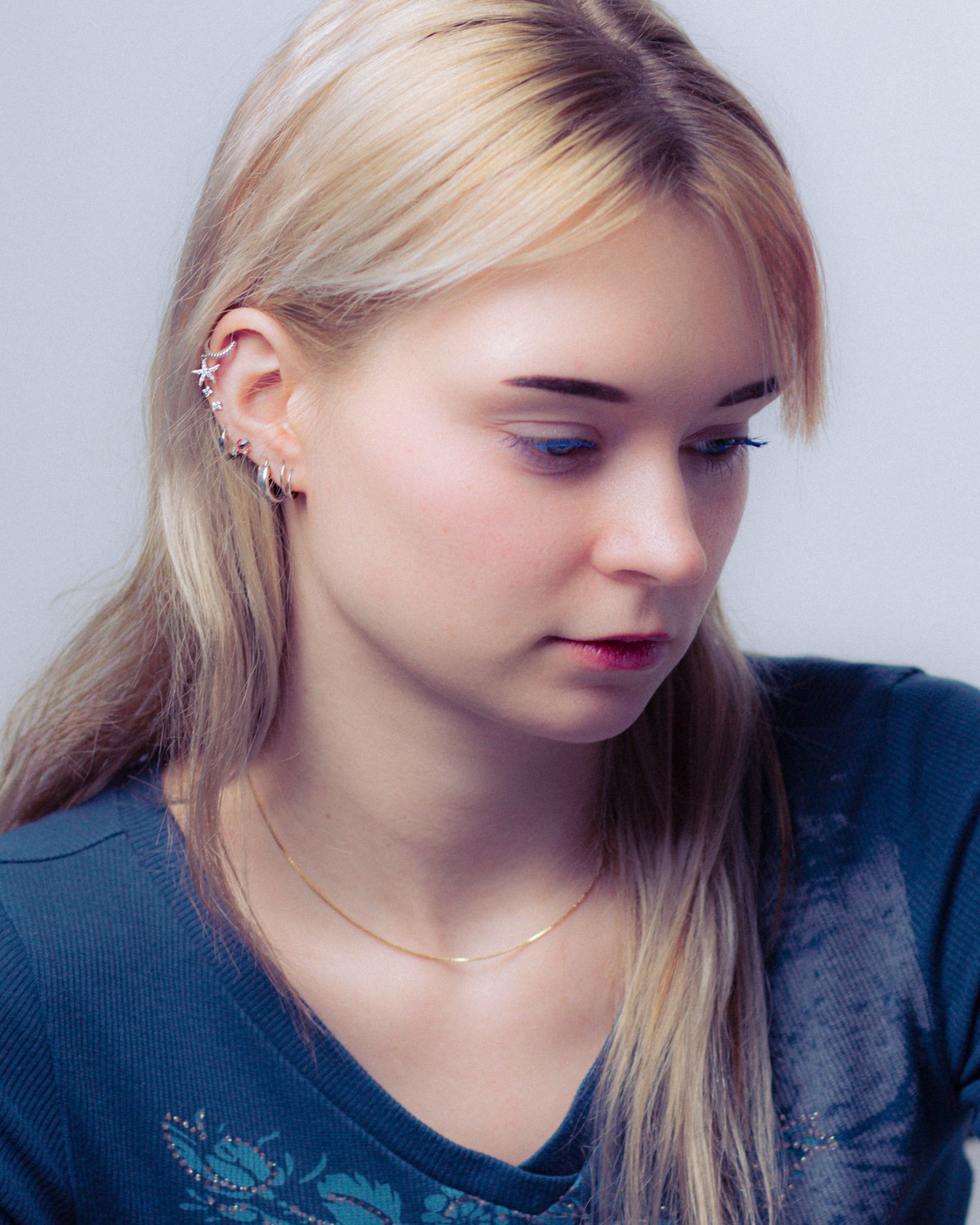 Young blonde woman with ear cuff jewelry and gold necklace wearing a blue top, looking downward.