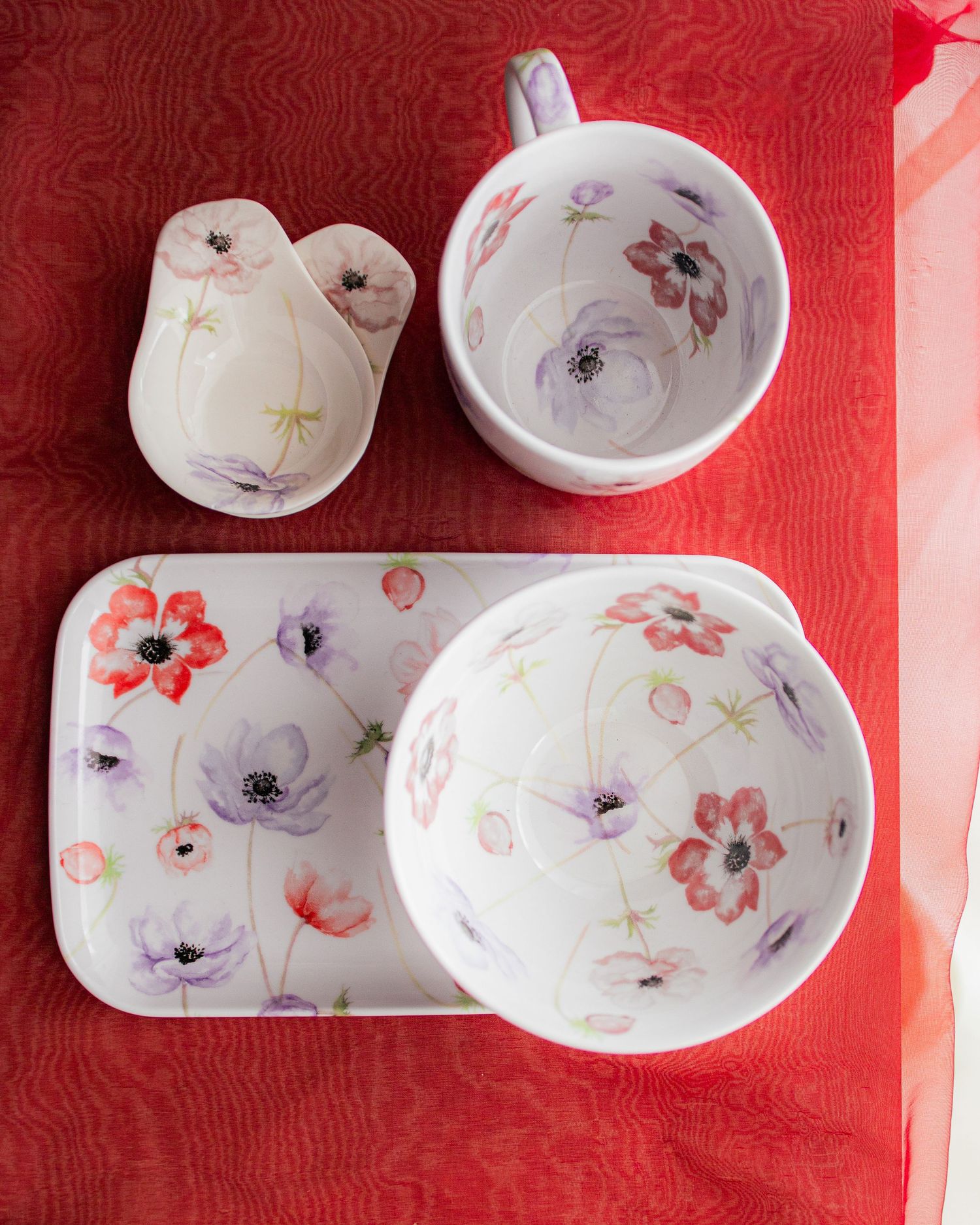White ceramic dinnerware set with colorful poppy floral pattern, including tray, bowl, mug, and small dish on red cloth.
