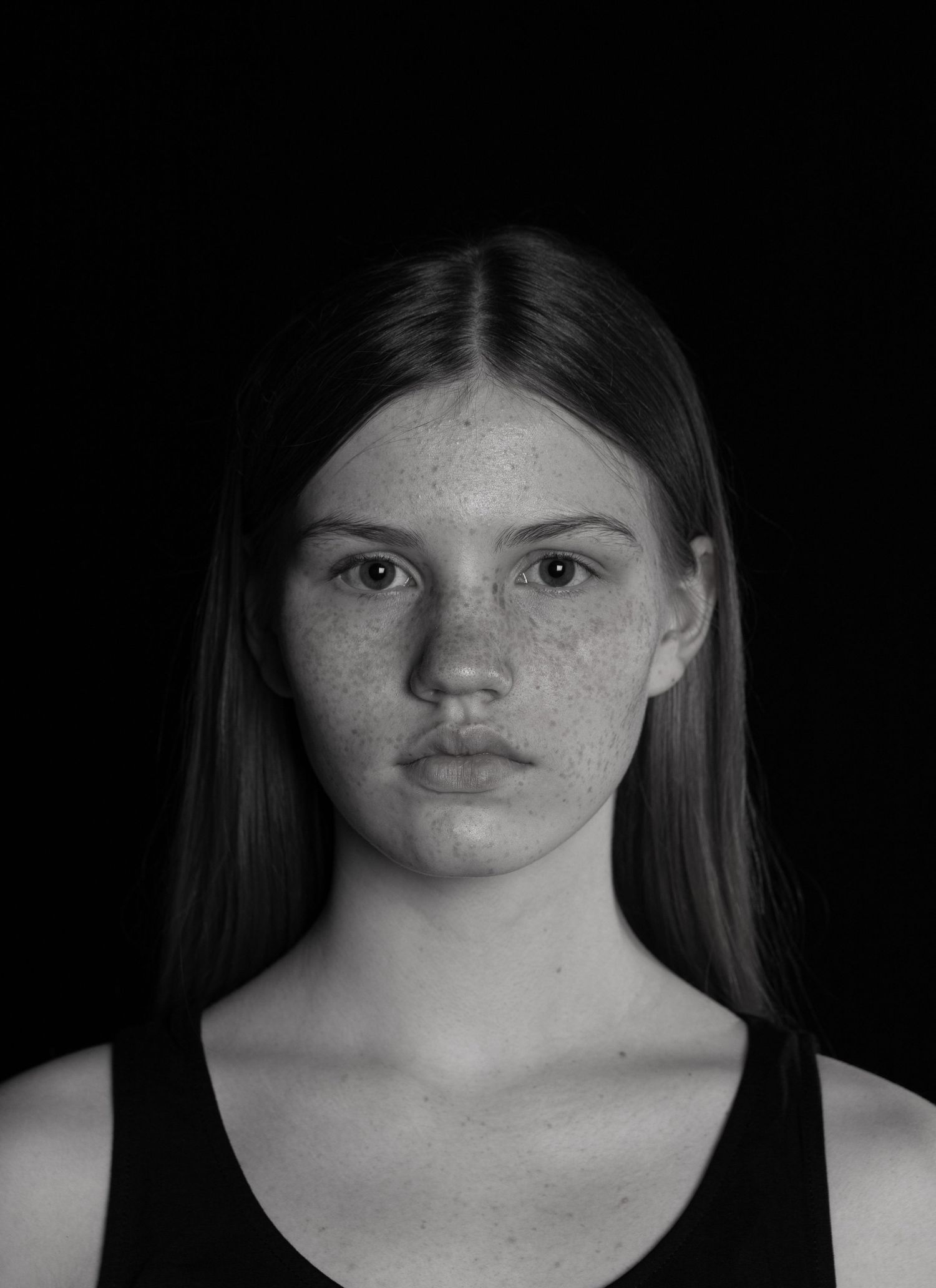 Black and white portrait of young freckled girl with long straight hair against dark background.