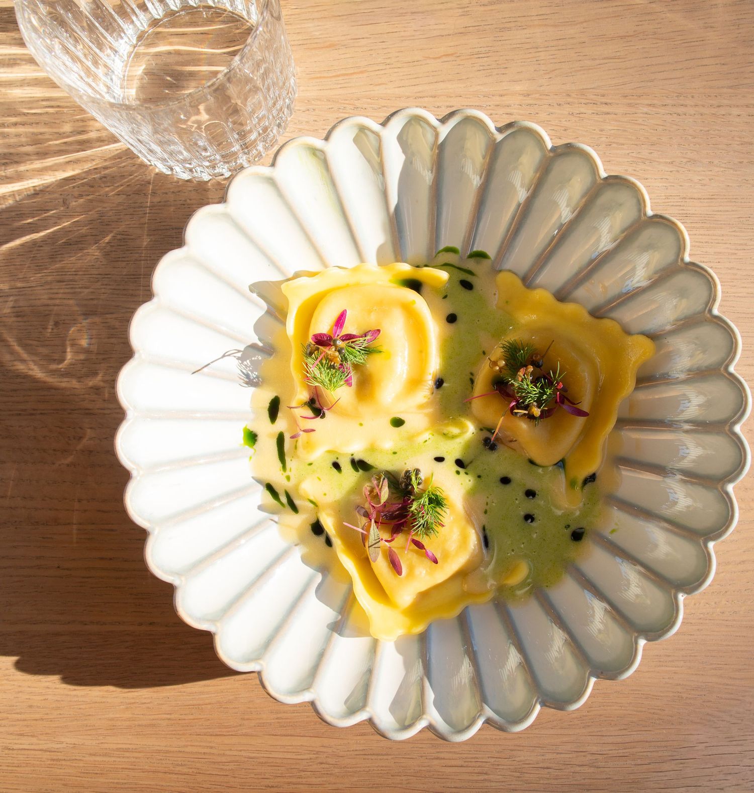 Golden ravioli pasta served with green herb sauce, black sesame seeds, and edible flowers on a white scalloped ceramic plate.