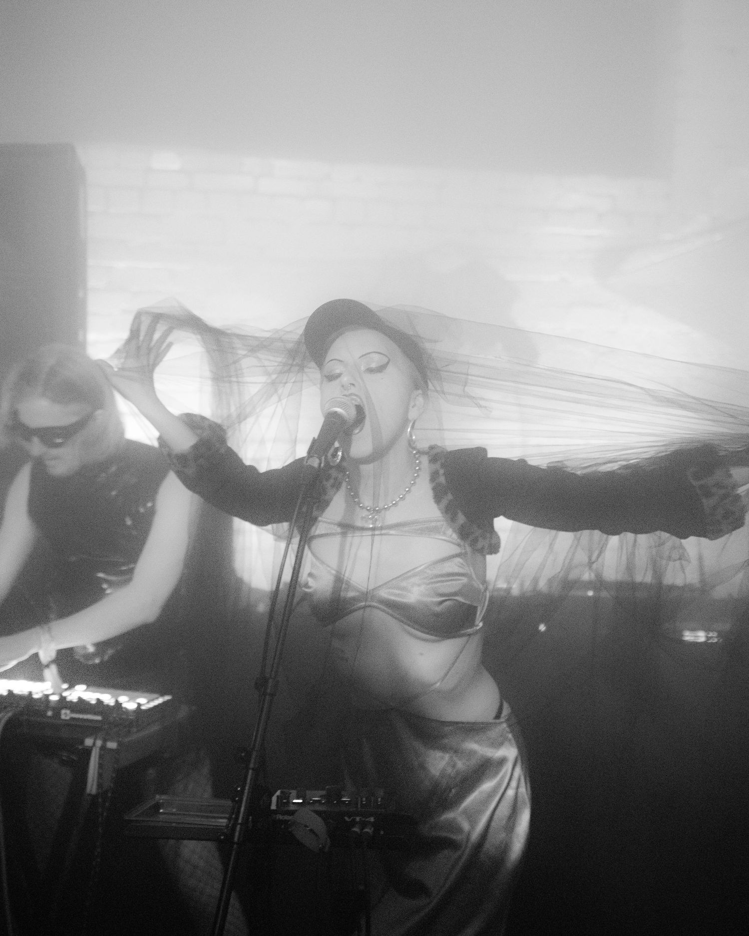 Black and white photo of a female singer performing on stage with a veil flowing behind her and a keyboardist in the background.