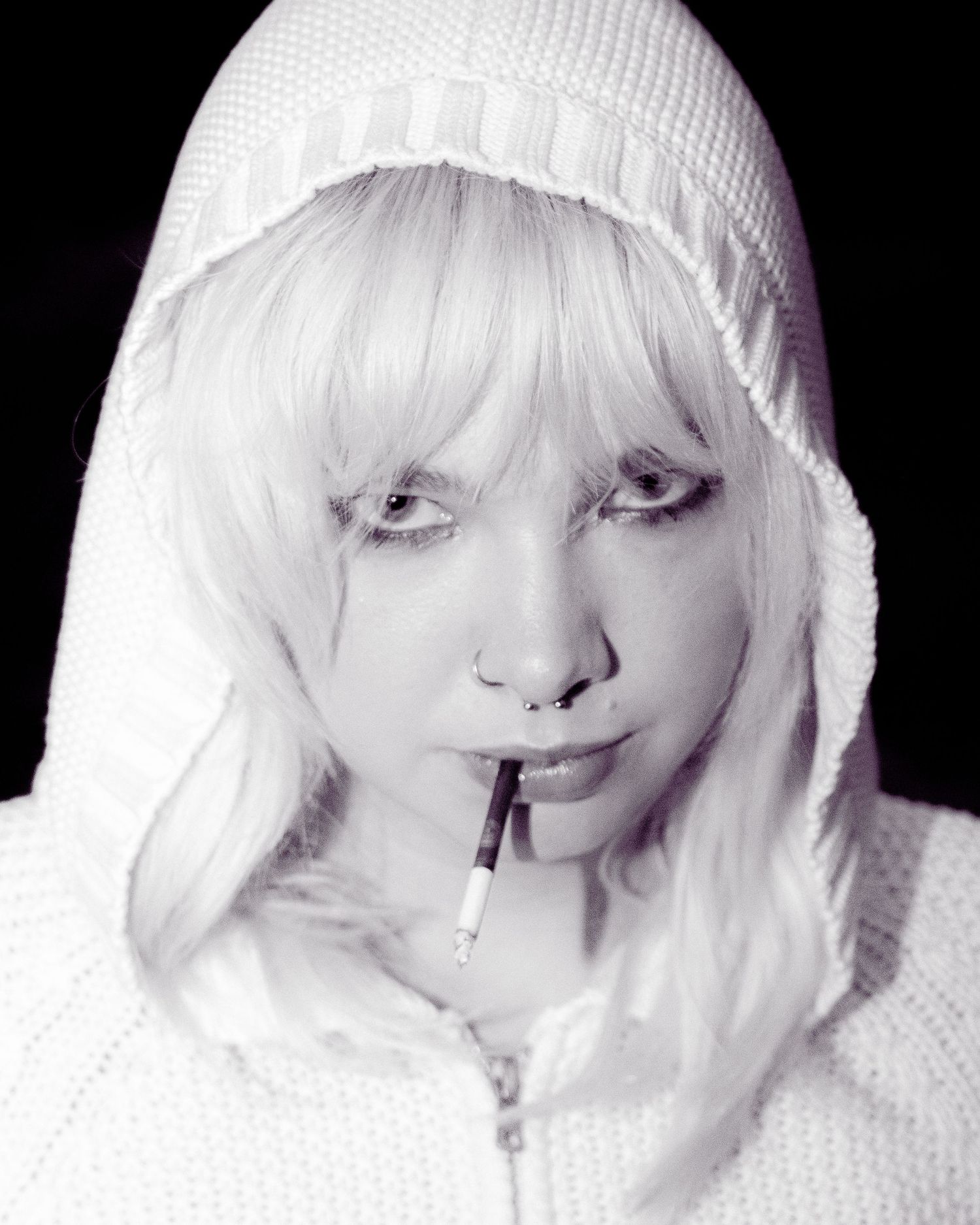Black and white portrait of woman in white hoodie with platinum hair holding cigarette in mouth.