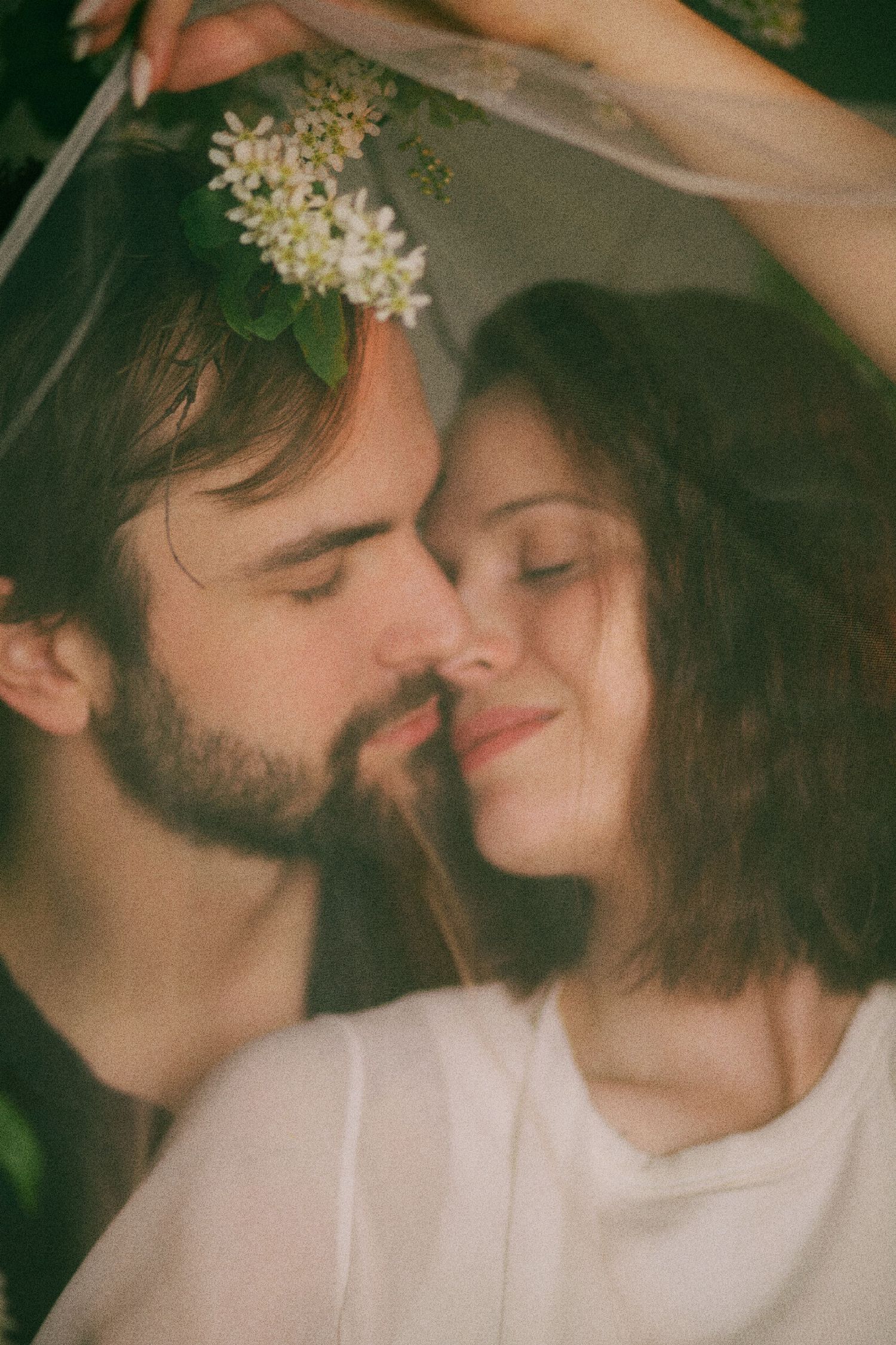 A romantic couple with closed eyes nearly kissing under a floral veil with white blooms in soft natural light.