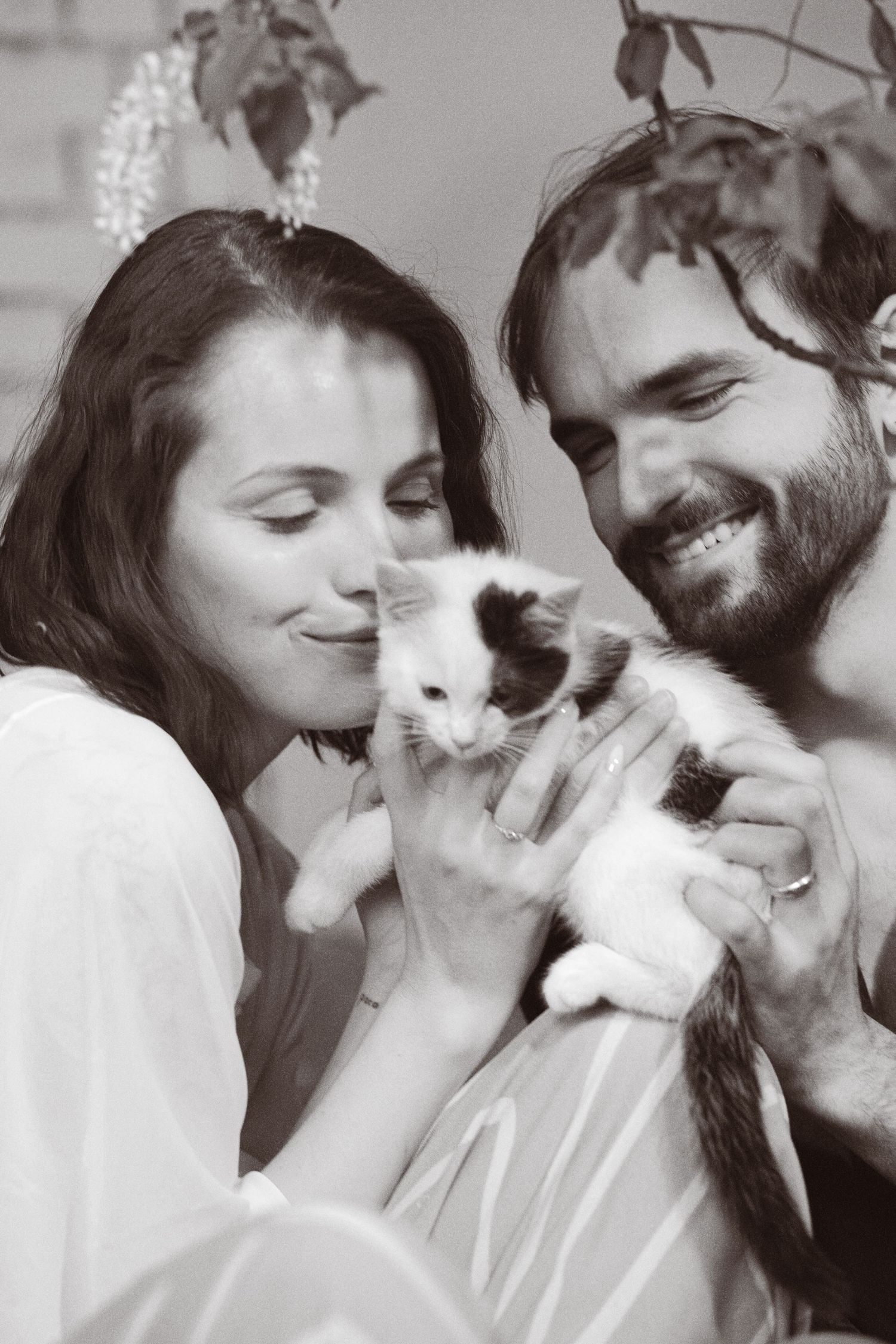 Black and white photo of a smiling couple tenderly holding a small kitten together near leafy plants.