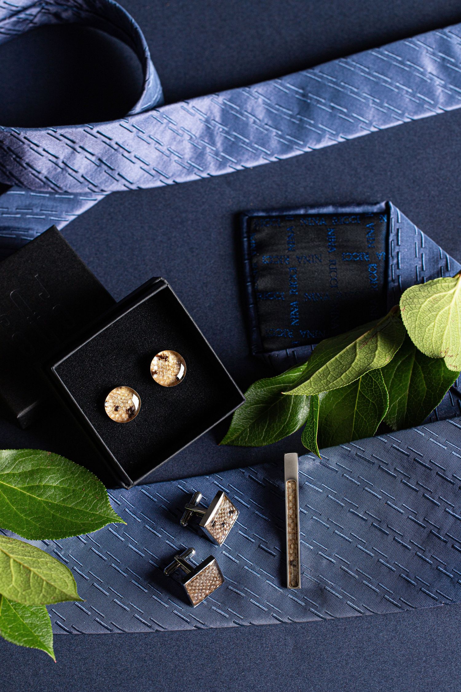 Gold round cufflinks in black box with blue striped tie and green leaves flatlay.