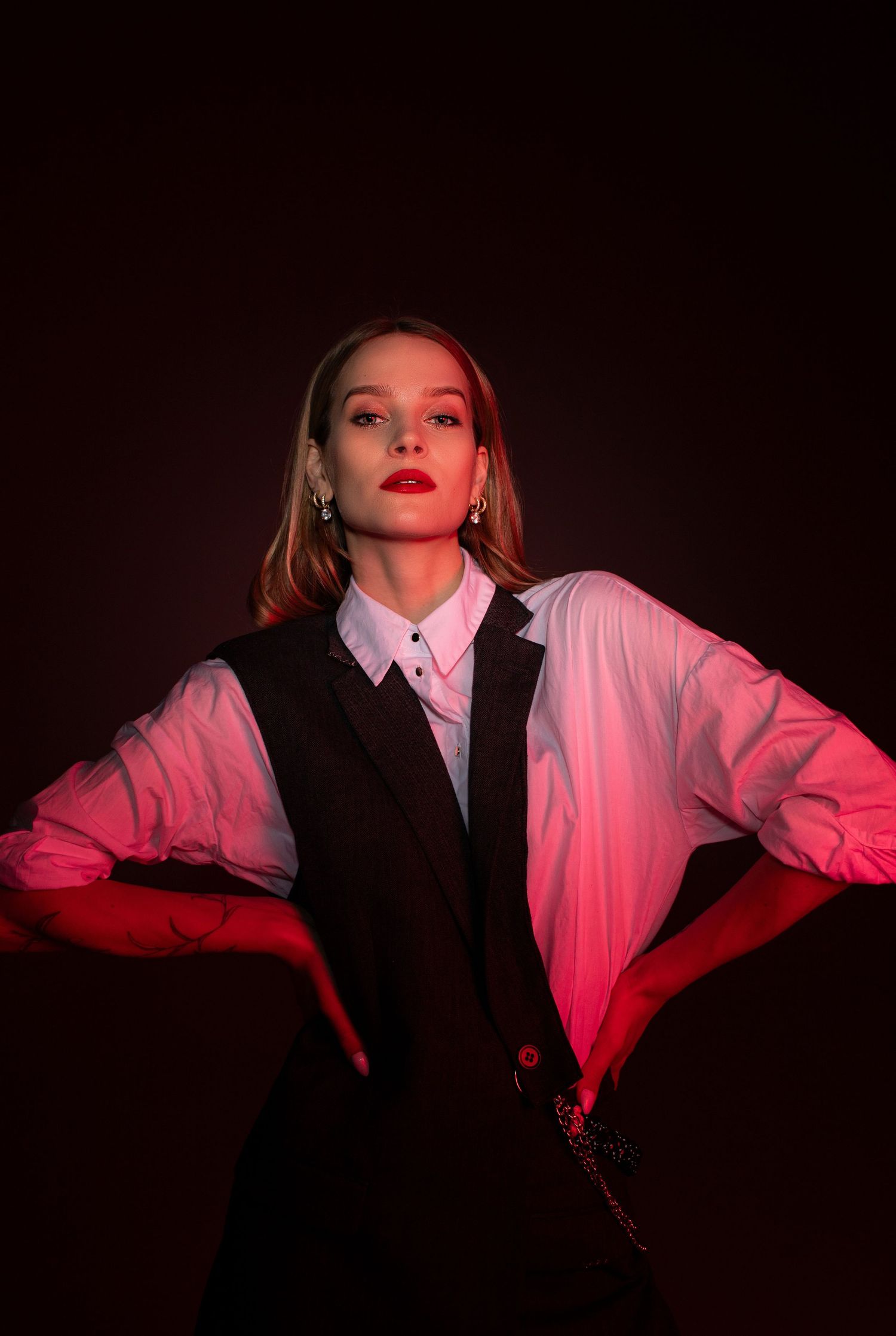 Stylish woman in black vest and white shirt posing with hands on hips under red studio lighting.