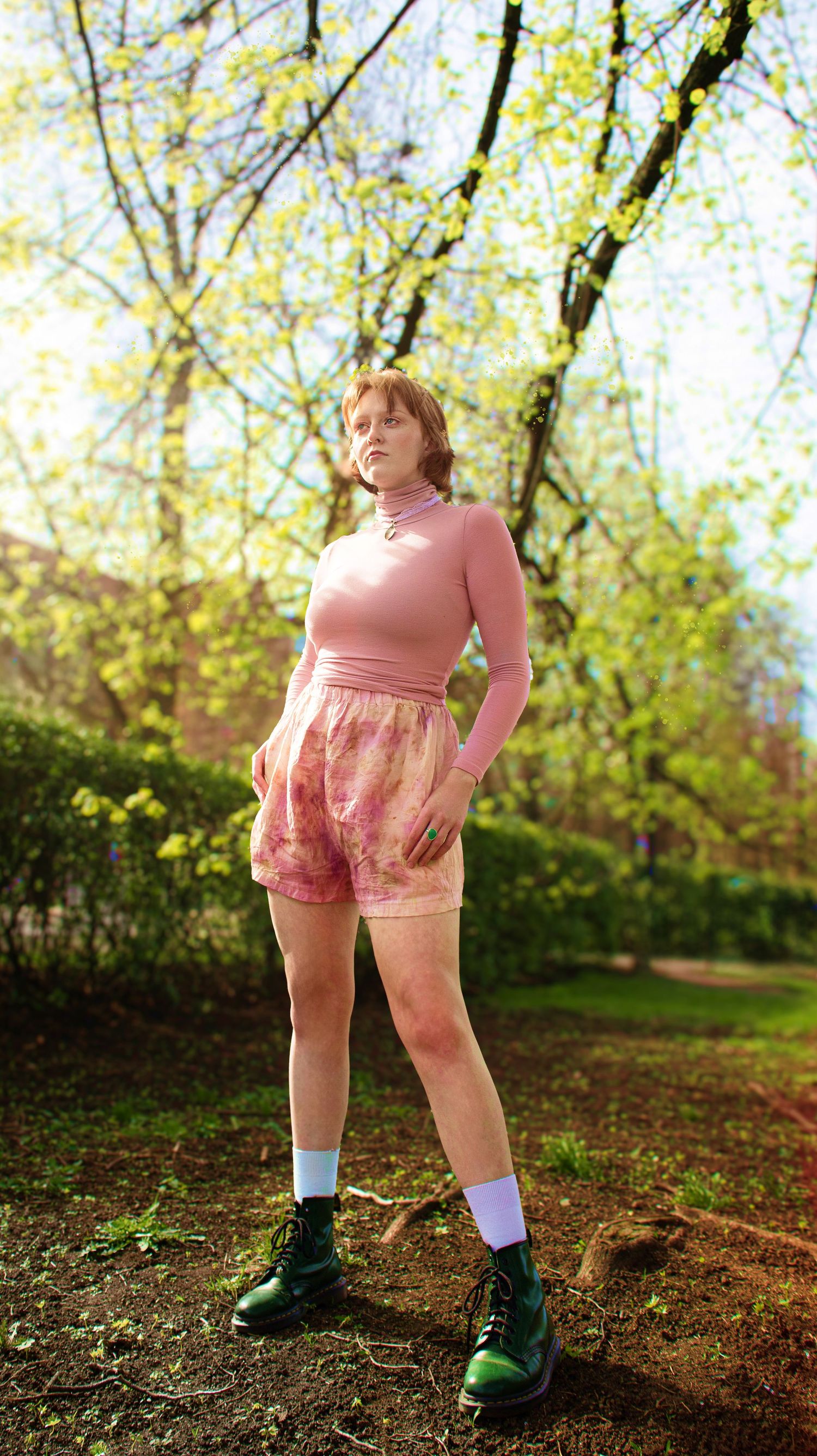 Young woman in pink turtleneck and tie-dye shorts posing outdoors among spring trees in green boots.