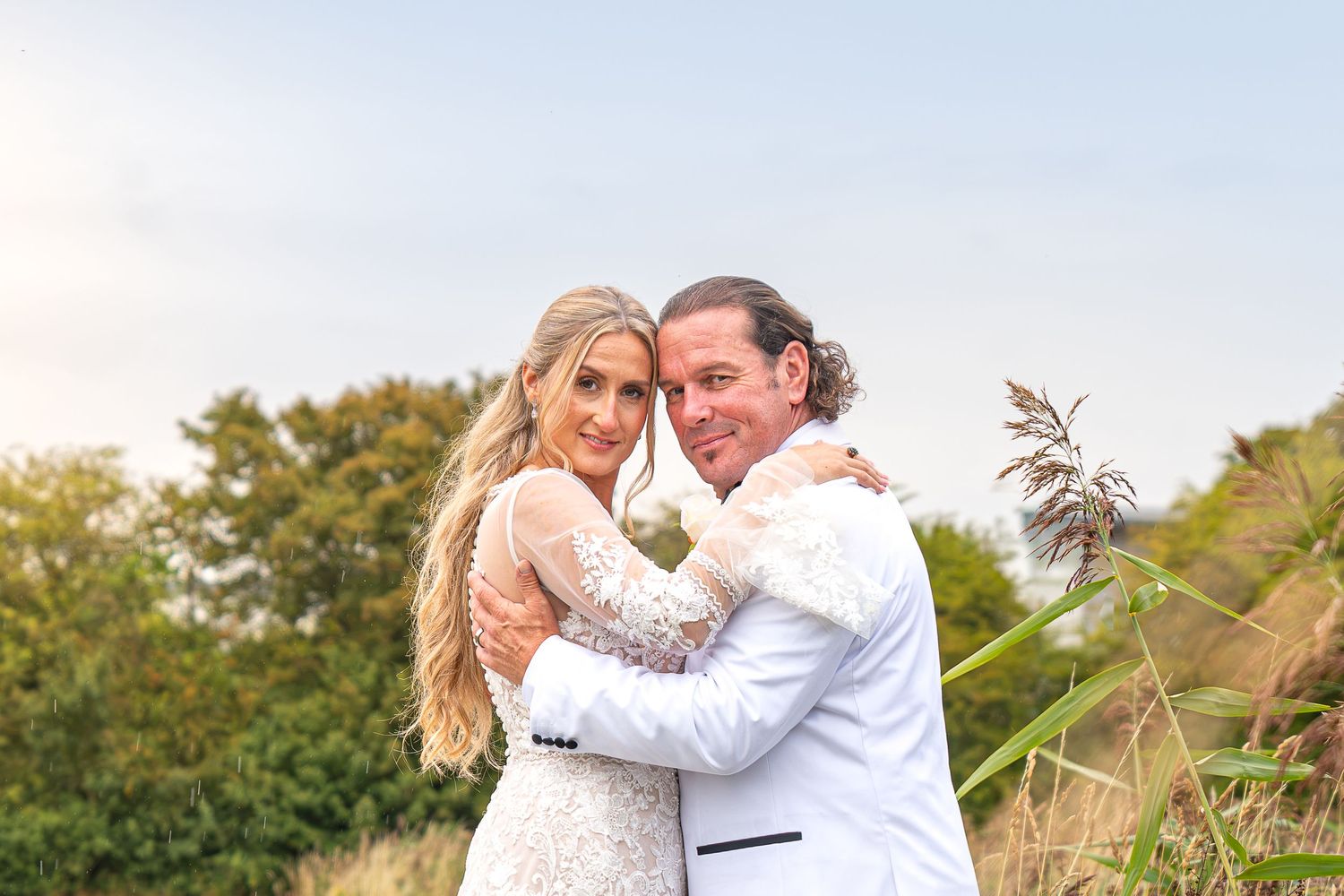 A bride and groom embrace outdoors in a lush garden setting, wearing elegant white wedding attire on their special day.