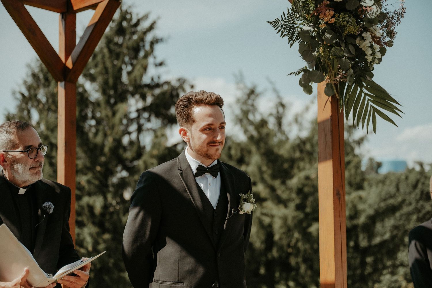 A couple in elegant wedding attire embraces during their outdoor ceremony under a wooden arch pergola. Pittsburgh Wedding Photographer