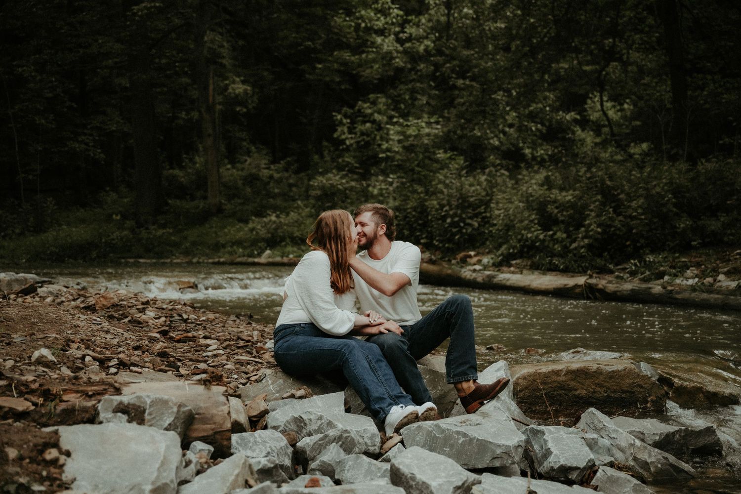 A romantic couple embraces on rocks by a river during an autumn engagement photoshoot in nature.