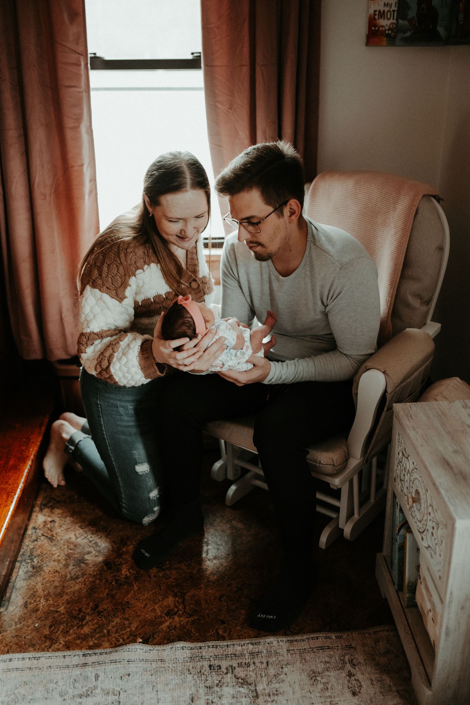 Two people sharing a tender moment while looking at their newborn in a cozy room with pink curtains. Pittsburgh / Westmoreland Photographer