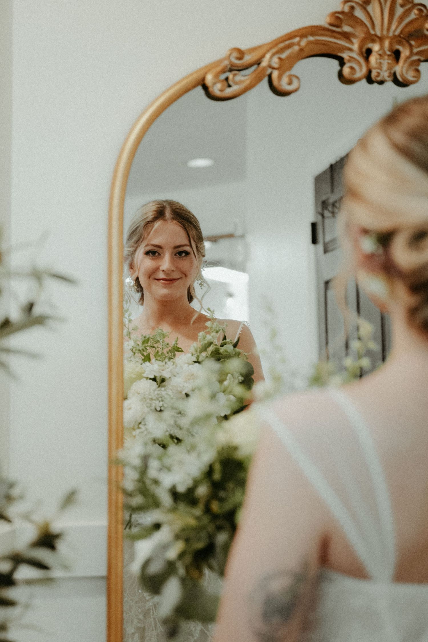 A reflection in an ornate gold mirror shows a person wearing a white wedding dress and holding a bouquet of flowers.