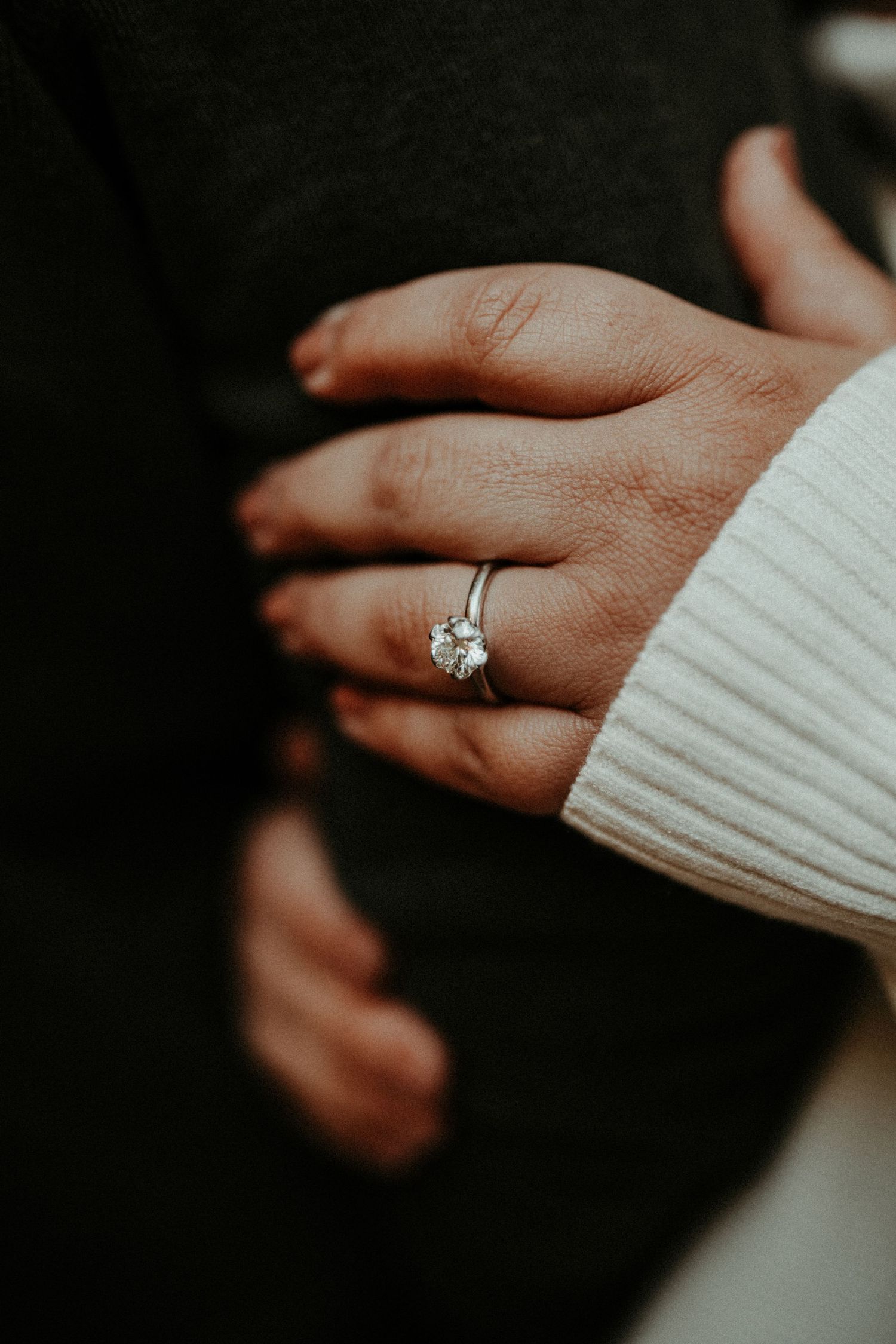 Close-up of diamond engagement ring on hand wearing white sweater against dark background. Pittsburgh Wedding Photographer