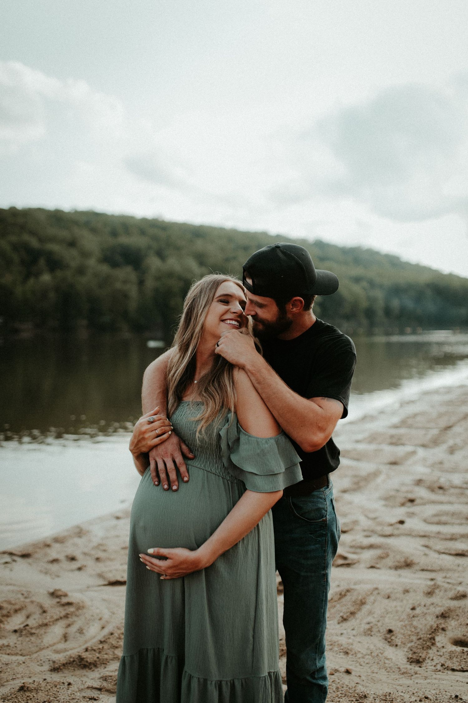 Romantic maternity photo of couple embracing on beach with lake and forest background. Pittsburgh Westmoreland Photographer