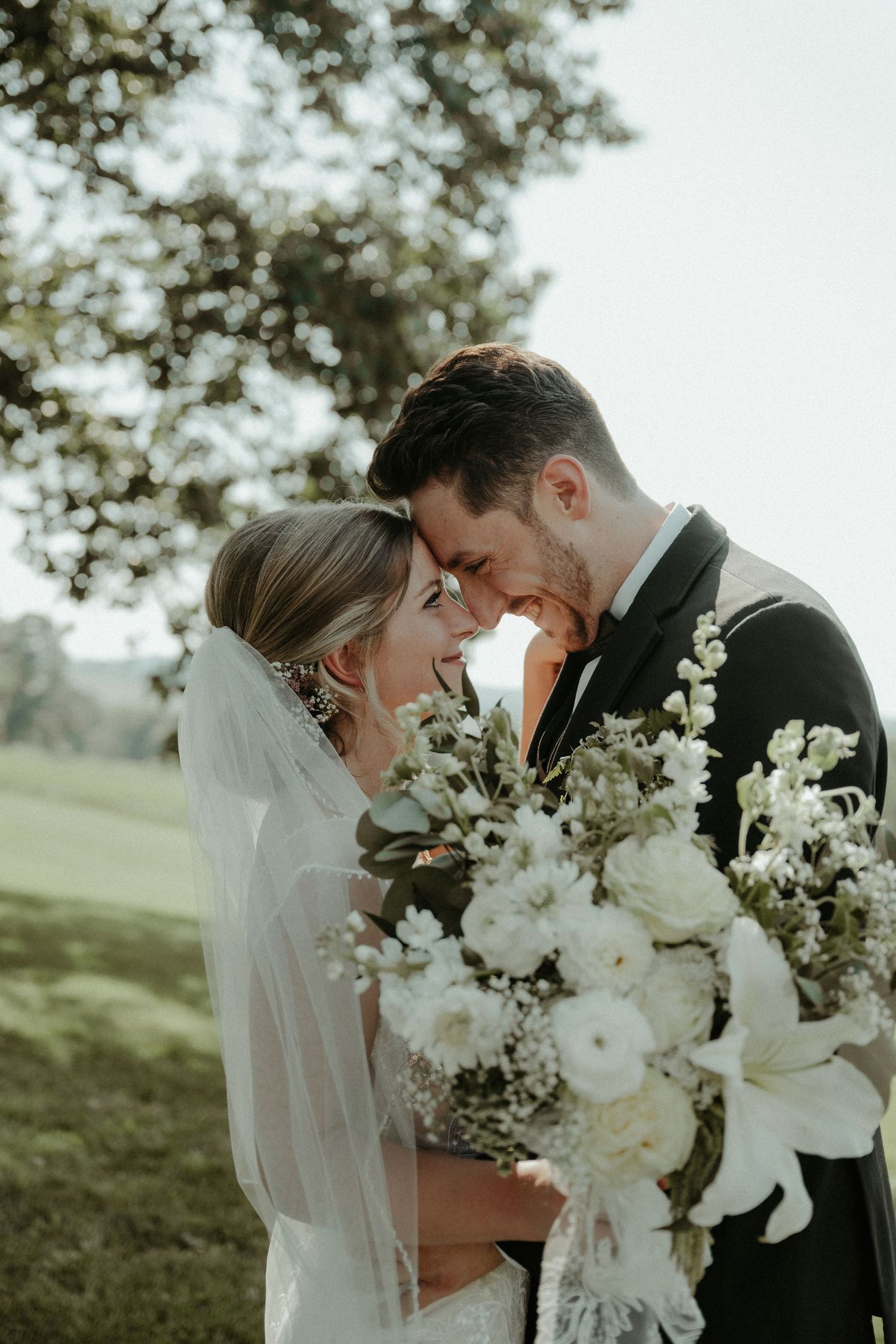 Romantic wedding couple embraces outdoors surrounded by white floral bouquets and ethereal greenery.