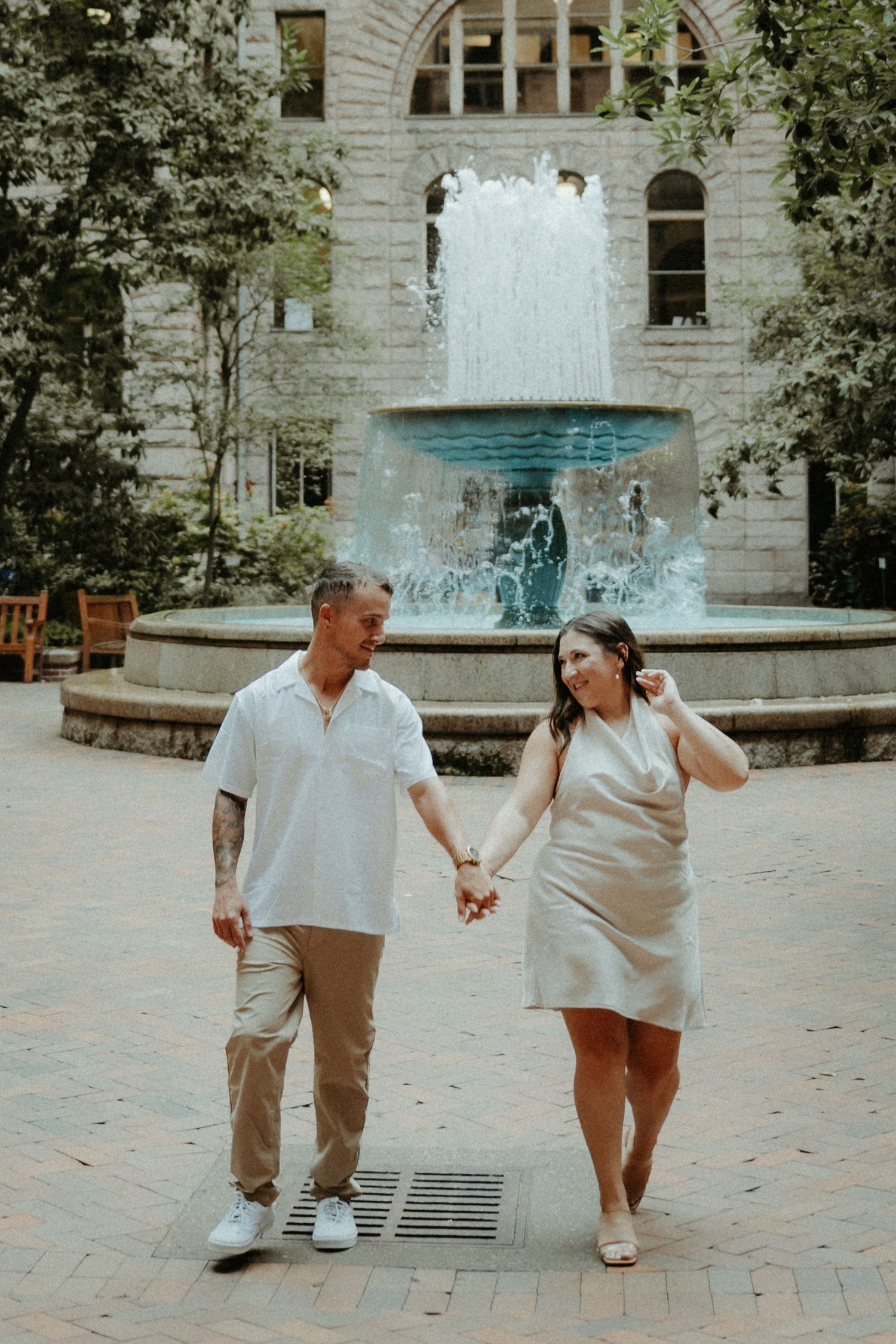 A couple walks hand in hand along a brick path near an ornate stone fountain on a warm sunny day. Pittsburgh Wedding Photographer