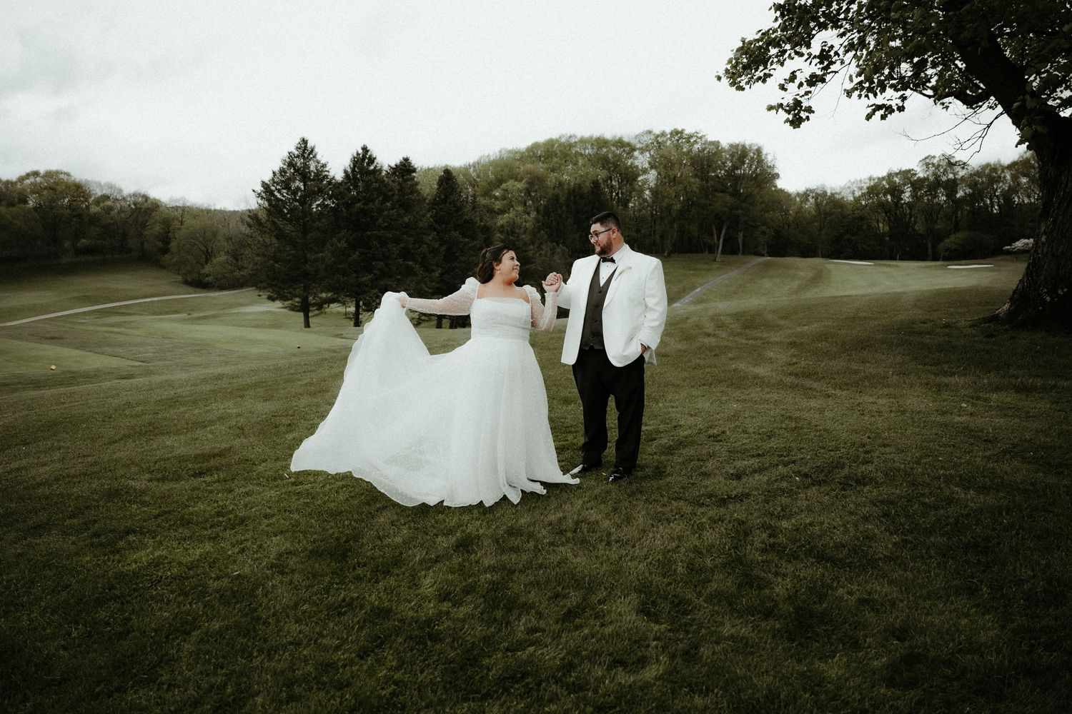 A couple in wedding attire stands together on a lush green golf course surrounded by trees on an overcast day.