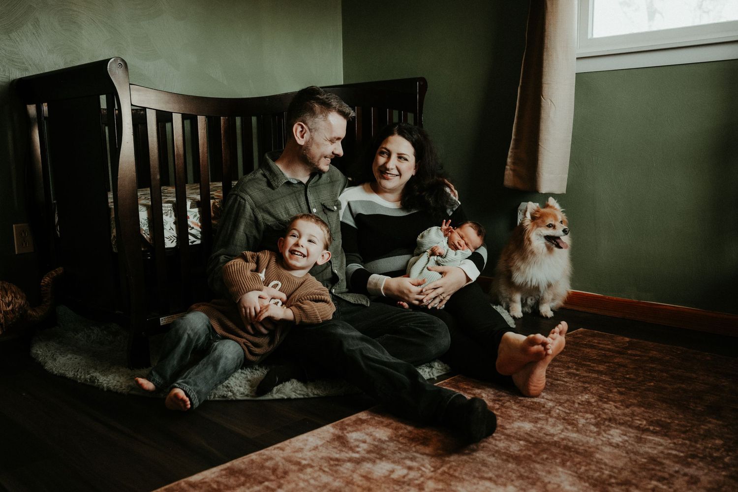 A family sits together on a wooden floor with children and their dog in a room with green walls. Pittsburgh / Westmoreland Photographer
