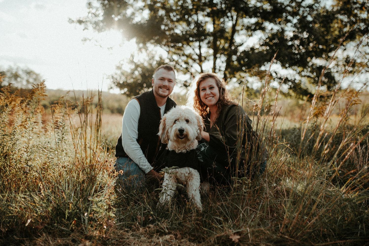 Two people kneel with their fluffy white Goldendoodle dog in a golden field during sunset.