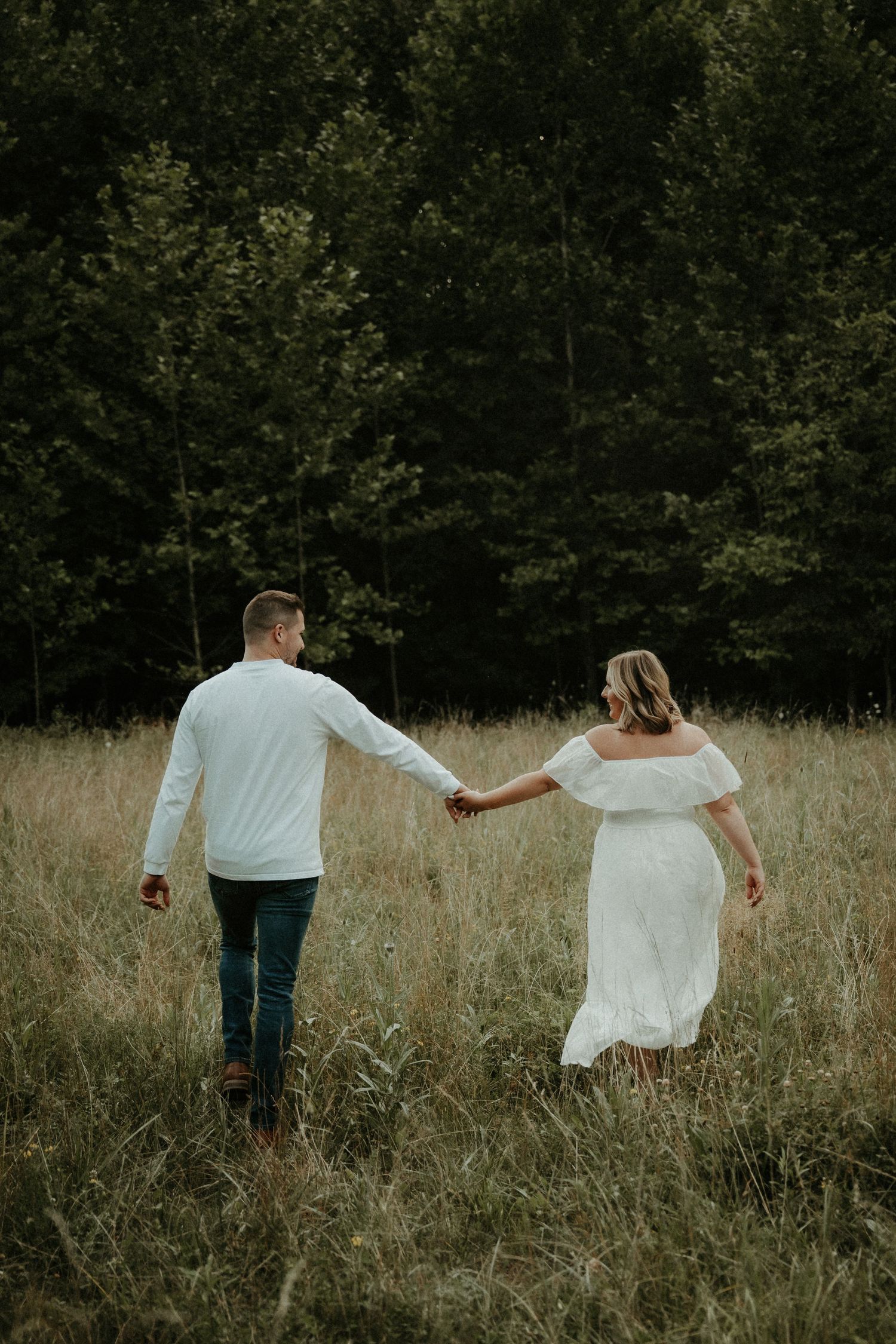 A couple holding hands walks through a tall grass meadow near a forest on a moody autumn day. Pittsburgh Wedding Photographer