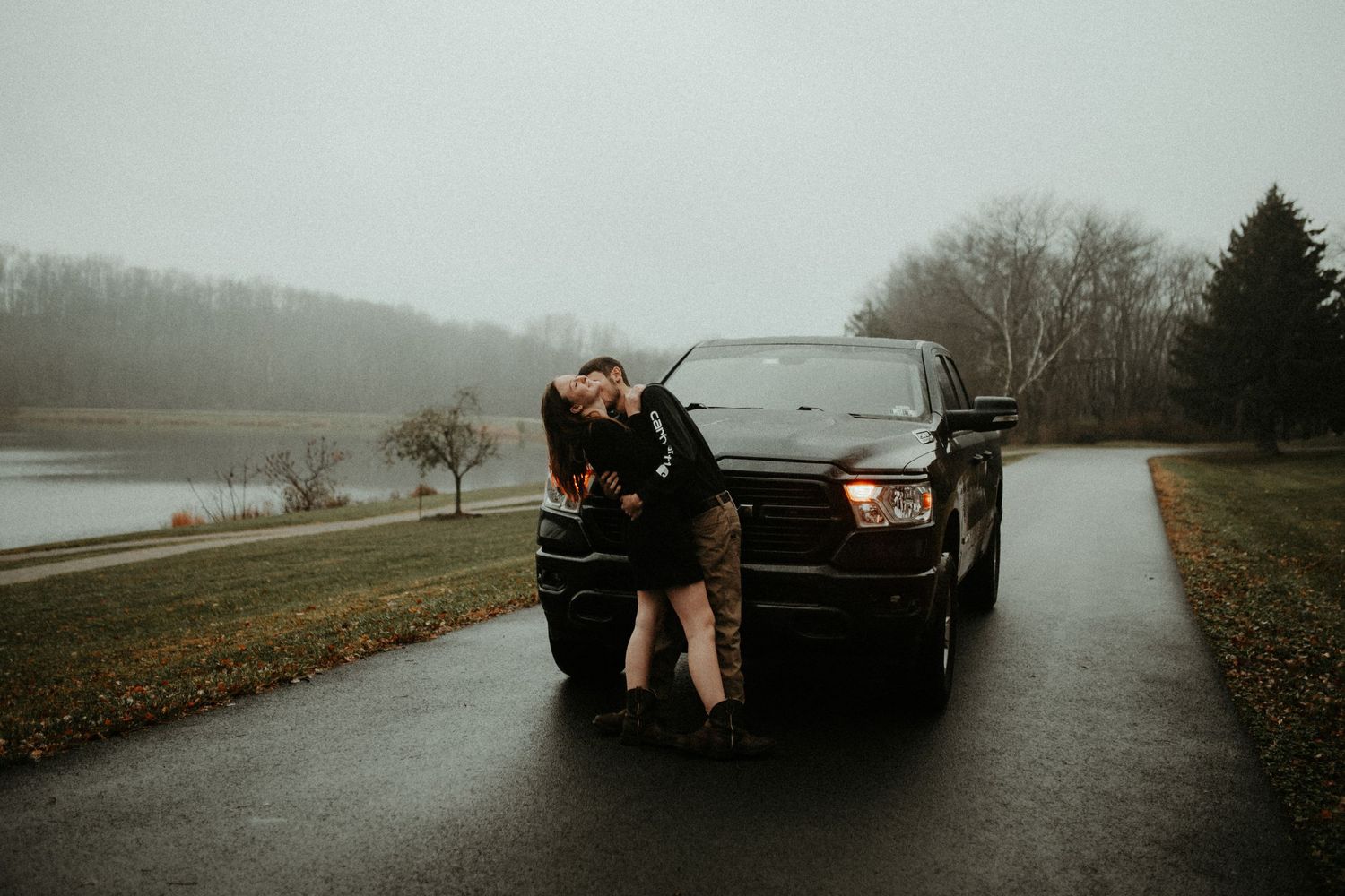 A couple embraces in front of a black pickup truck on a rainy day near a lake. Pittsburgh Wedding Photographer