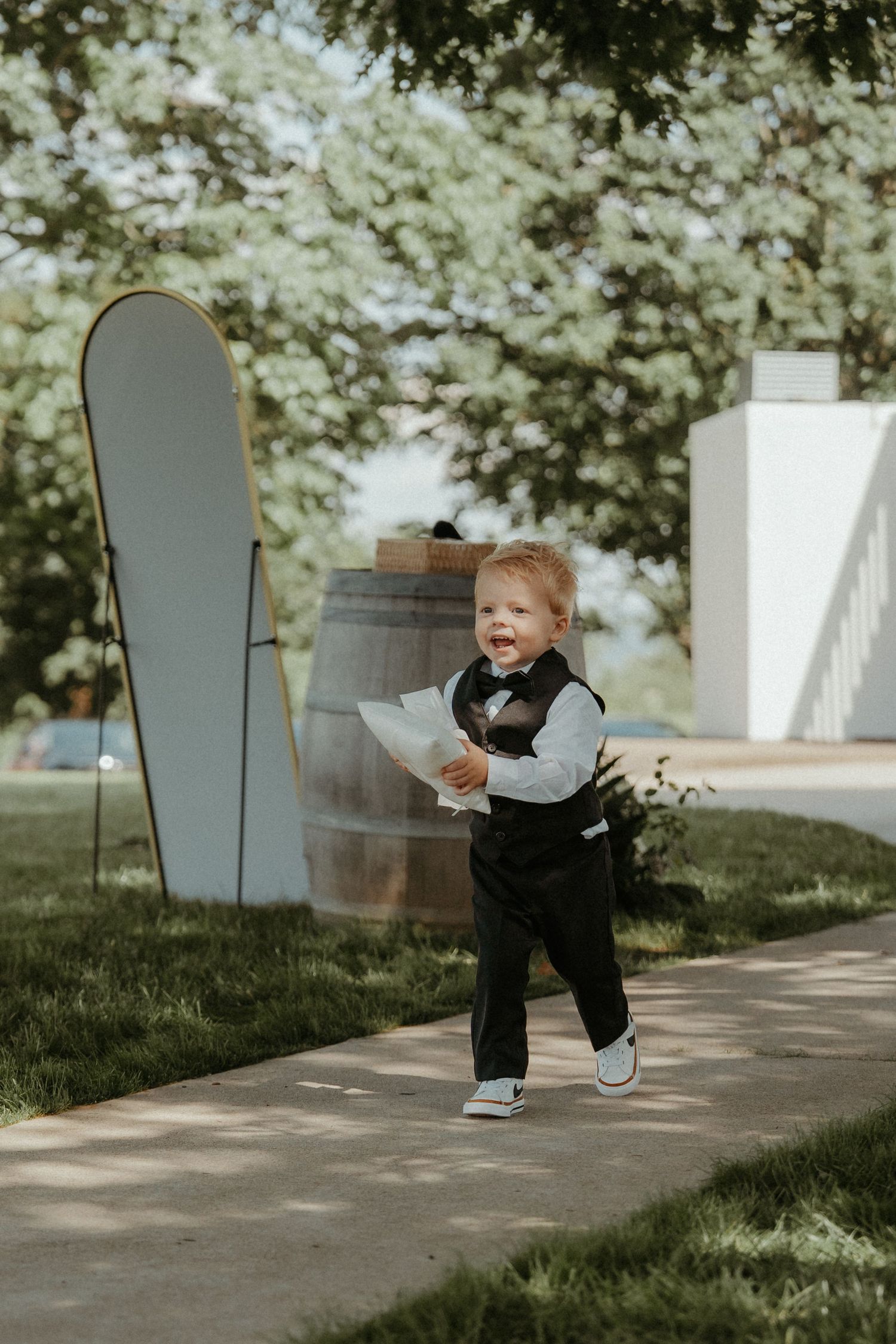 Small child in formal vest outfit plays near white markers and wooden barrel in garden setting.