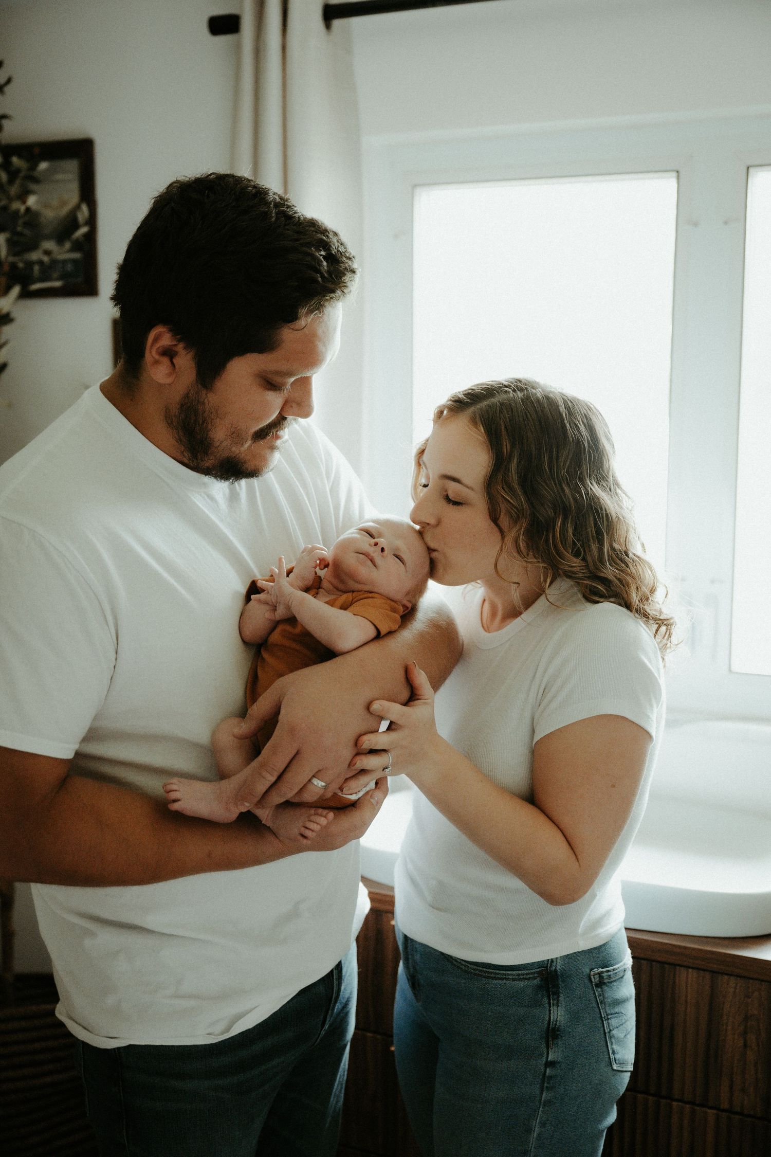 A couple shares a tender moment with their newborn baby in a bright, sunlit room. Pittsburgh / Westmoreland Photographer