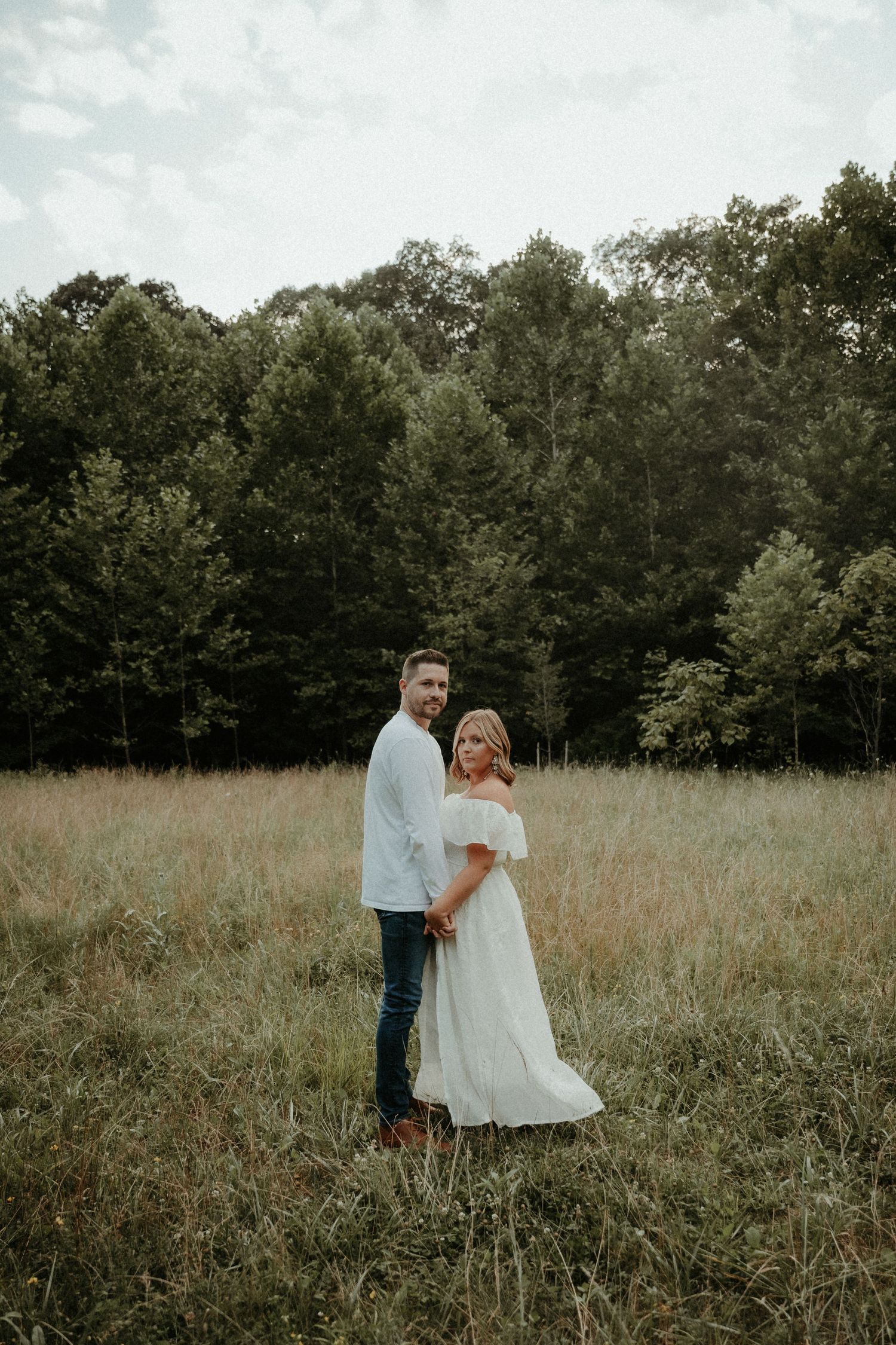 A couple poses together in a serene meadow with a forest backdrop during golden hour.