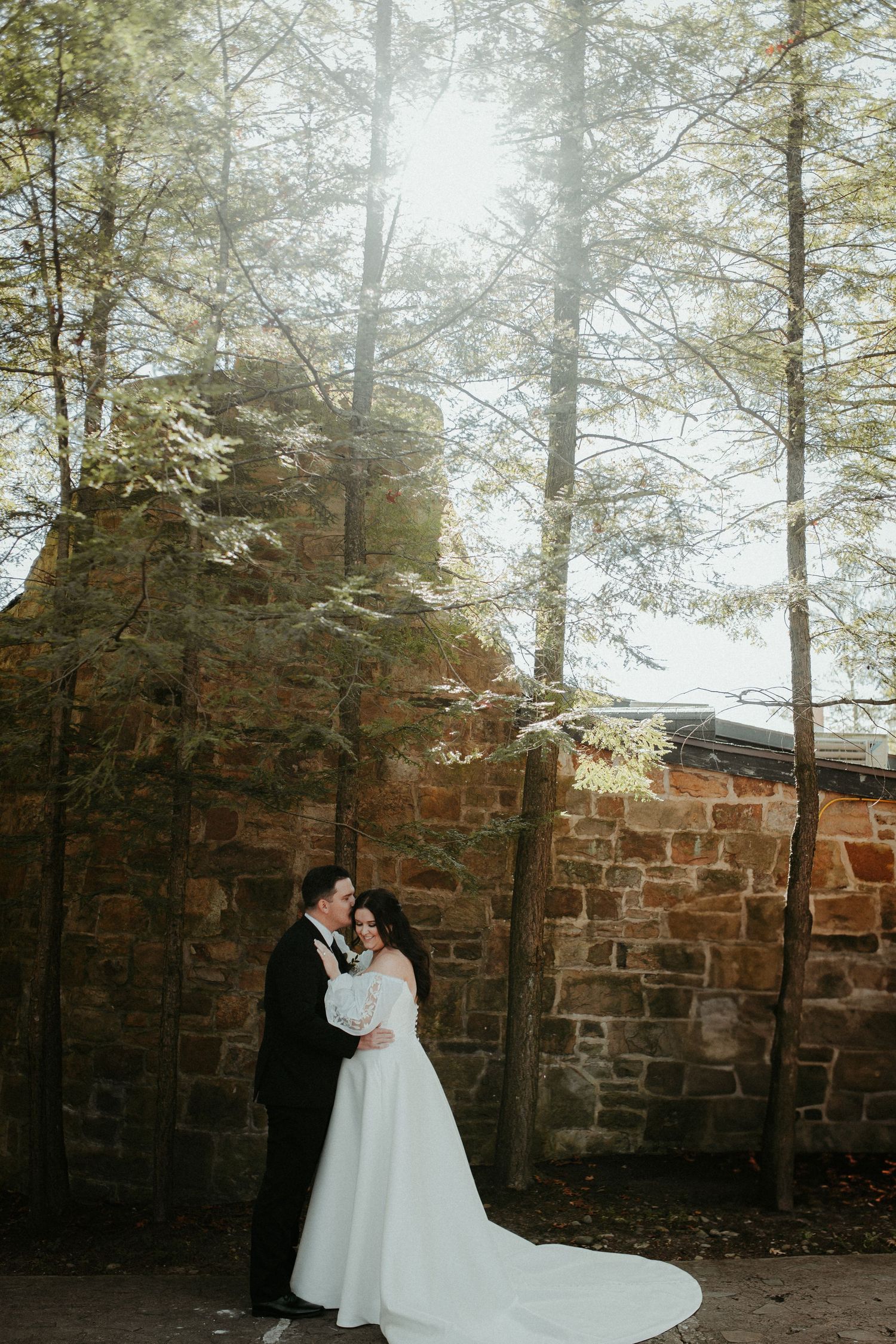 Newlyweds embrace against brick wall with sunlight filtering through trees. Pittsburgh Wedding Photographer