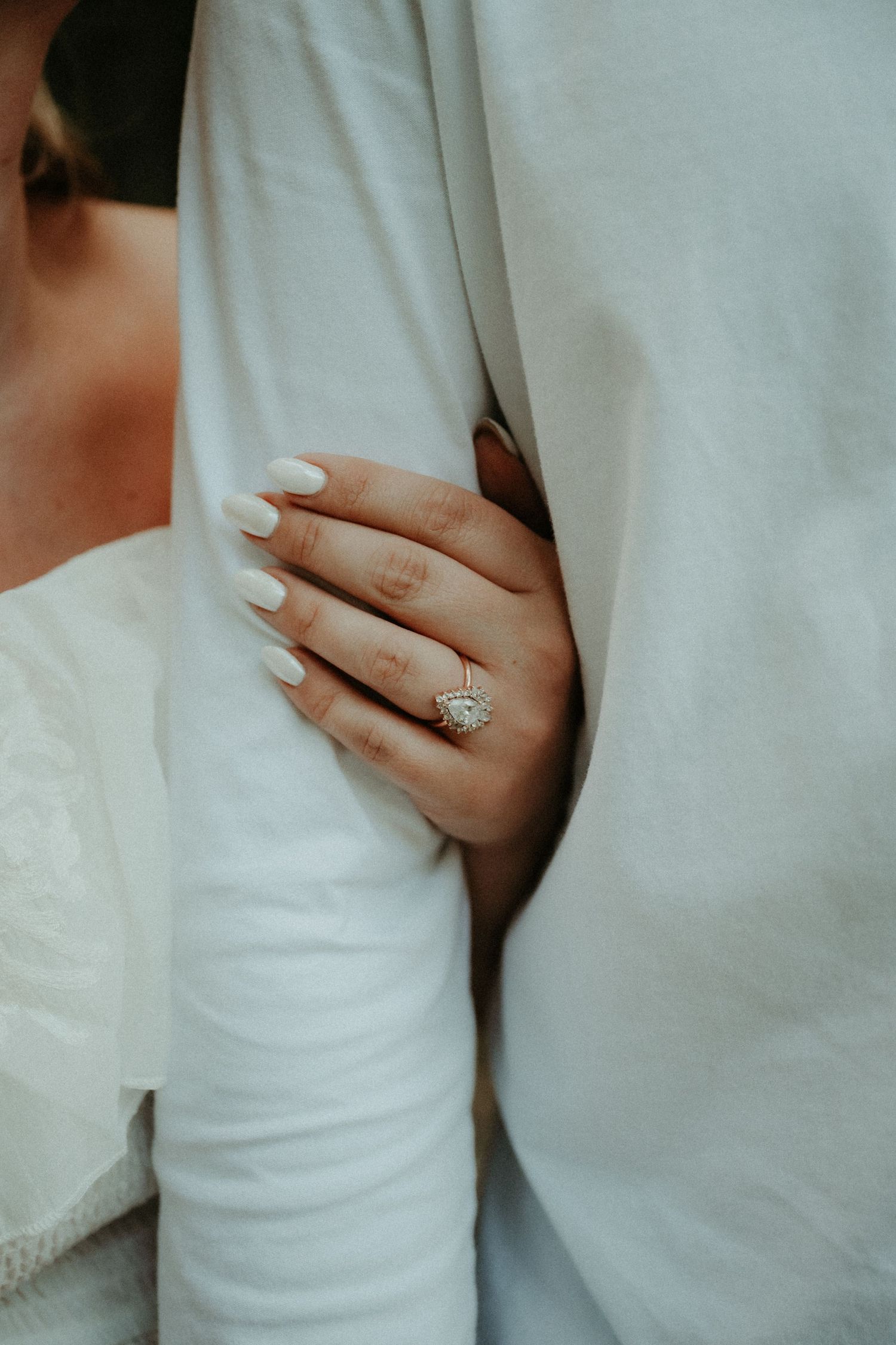 A close-up of a hand with white manicured nails wearing a sparkling diamond engagement ring against white fabric.