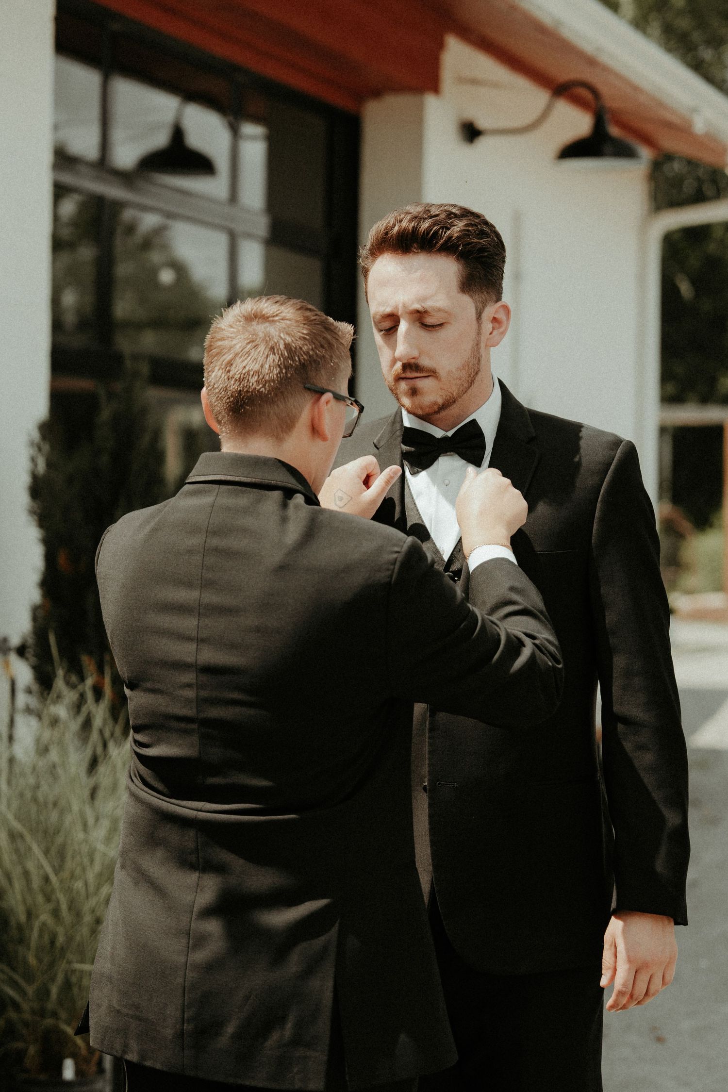 Wedding party members help adjust a groom's bowtie in a stylish black and white ensemble on a porch. Pittsburgh Wedding Photographer