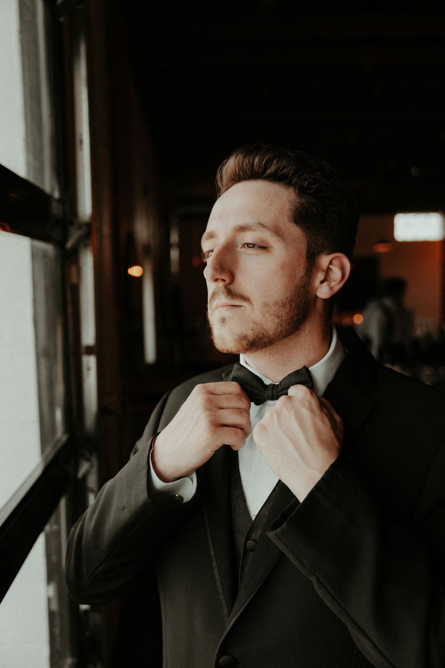 Moody portrait of someone adjusting a bowtie while wearing a formal black tuxedo near a window at dusk. Pittsburgh Wedding Photographer