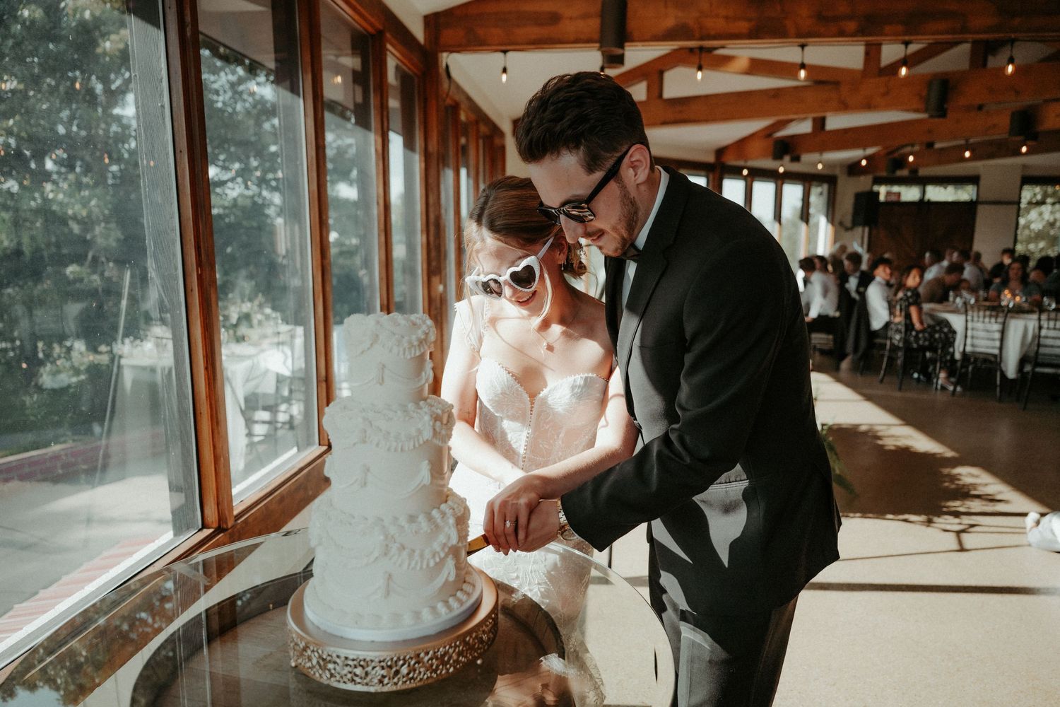 A bride and groom cut their elegant white wedding cake together in a sunlit venue with floor-to-ceiling windows.