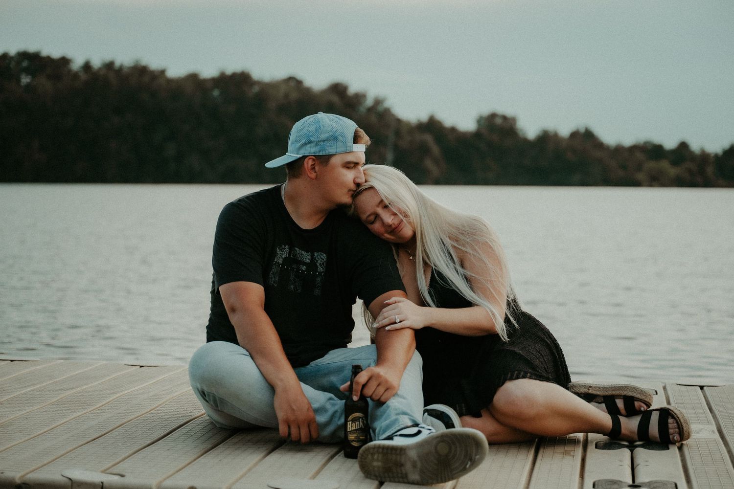 Two people sit together on a wooden dock by a scenic lake at sunset, creating a romantic silhouette.