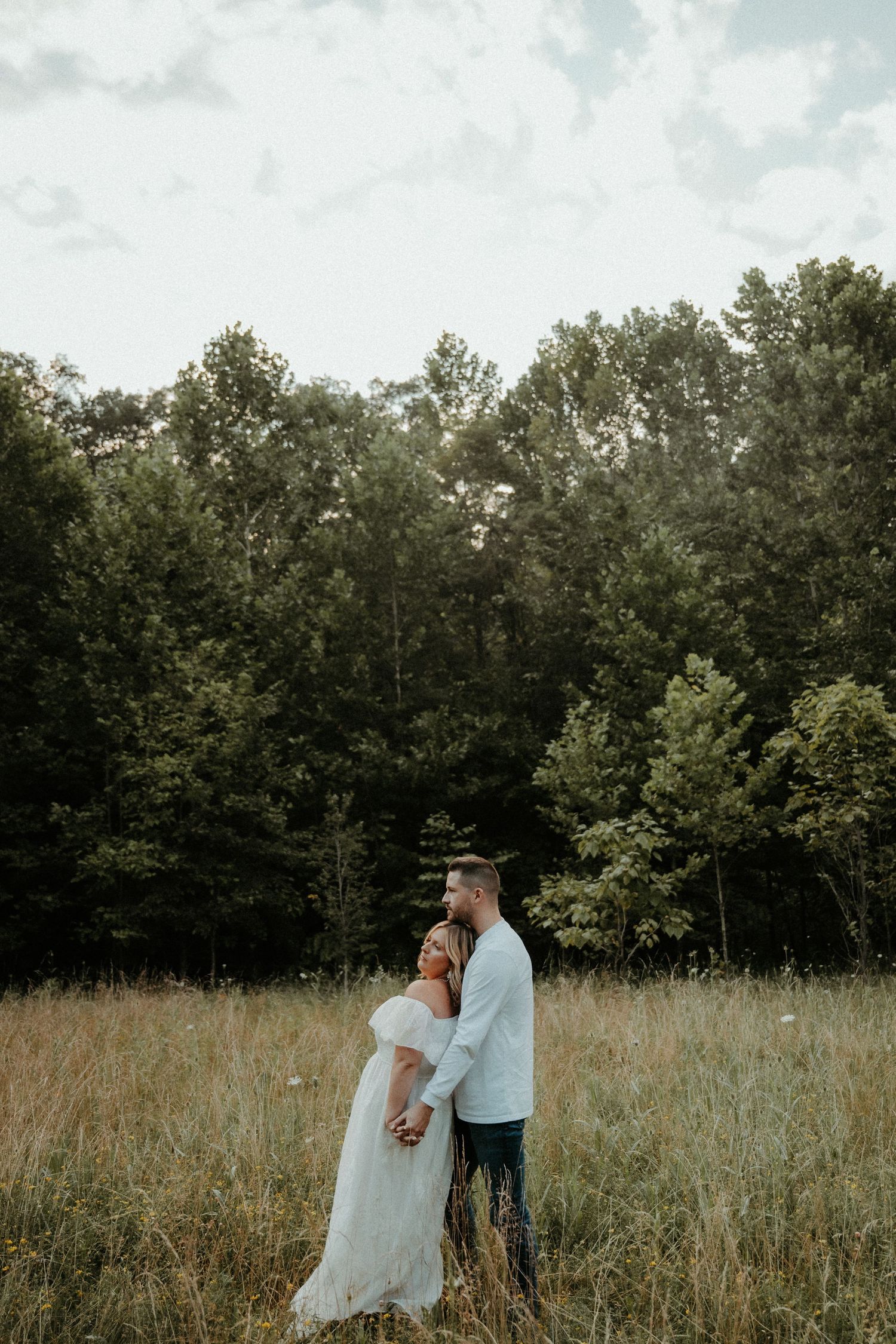 A couple in wedding attire embrace in a rustic field with trees in the background during golden hour.