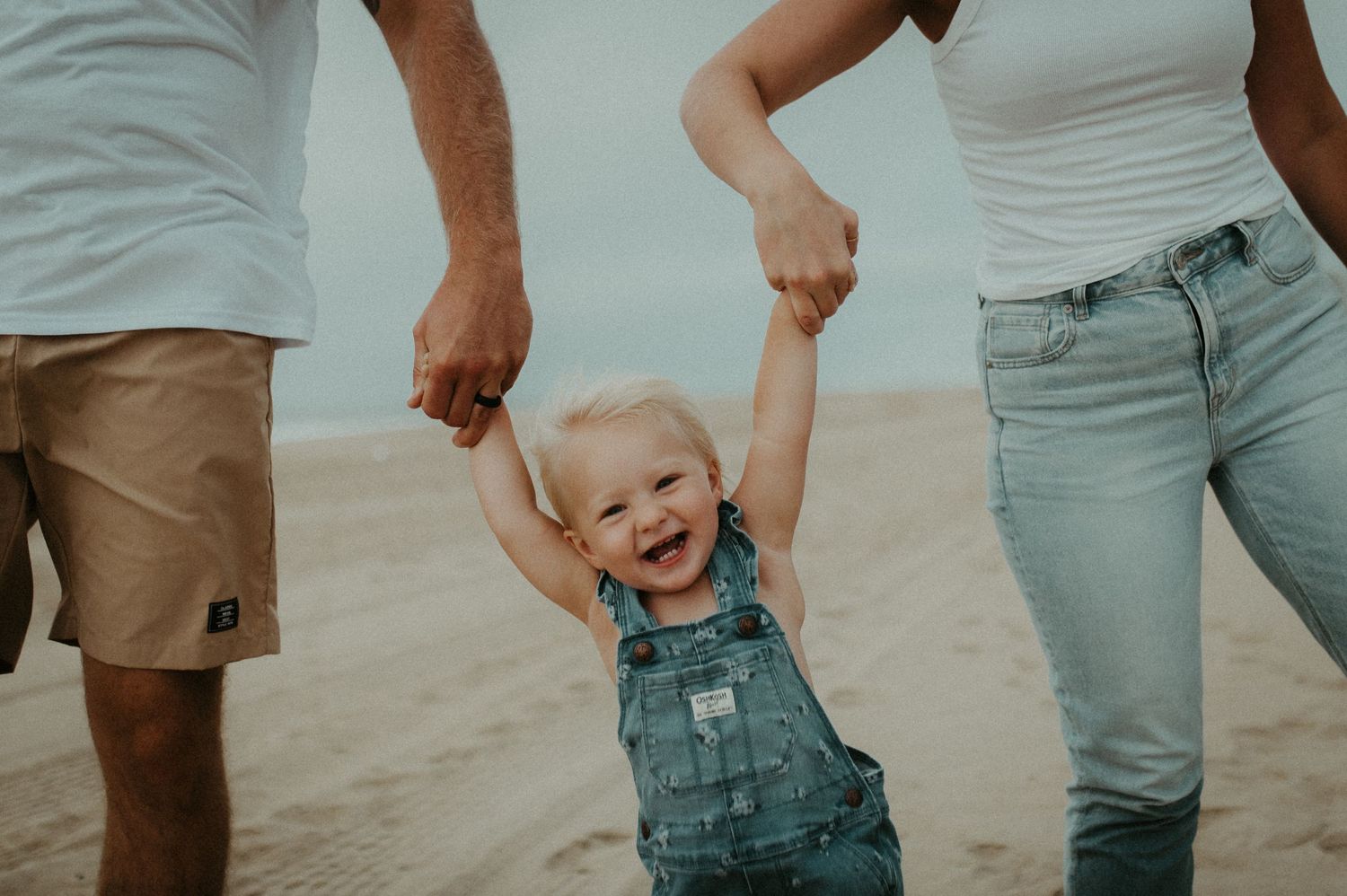 A joyful toddler in overalls swings between their parents on a sandy beach.