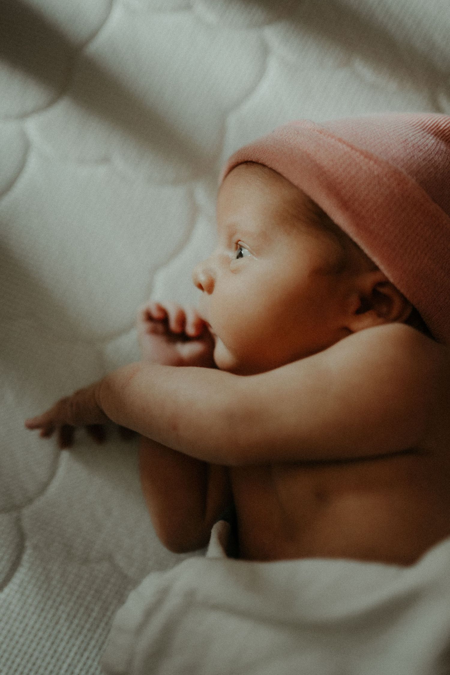A sleeping baby in a pink cap rests on white bedding in soft lighting with shadows cast across their gentle features.