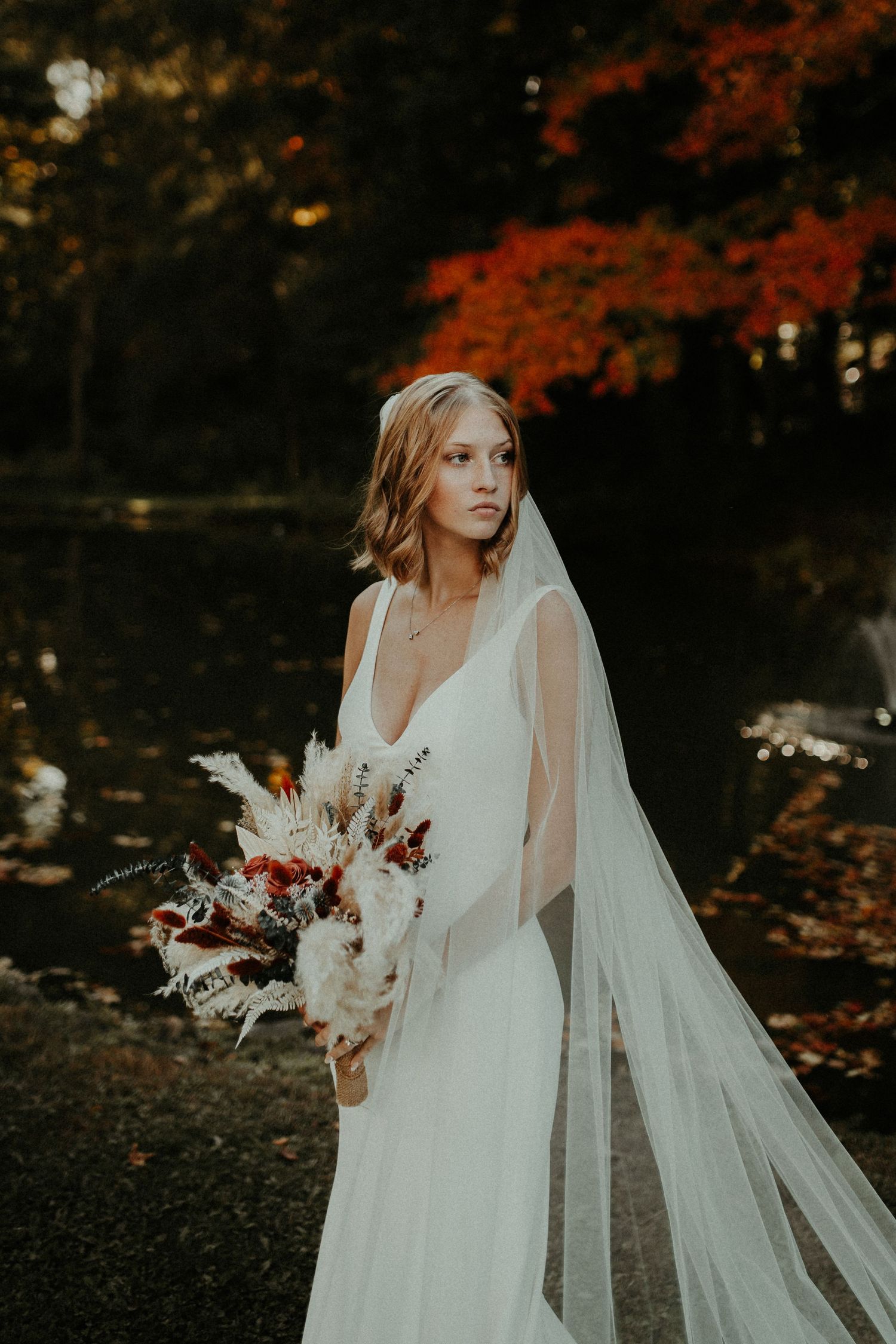 A bride in an elegant white gown holds a bouquet against autumn foliage at sunset. Pittsburgh Wedding Photographer