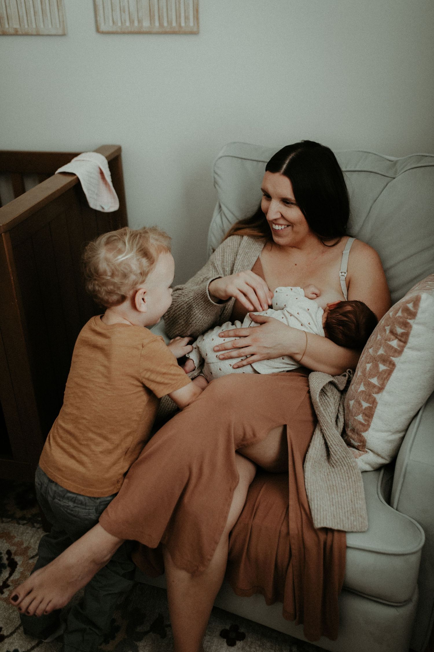 A heartwarming family moment as a woman in rust-colored clothing sits on a couch nursing an infant while a toddler looks on.
