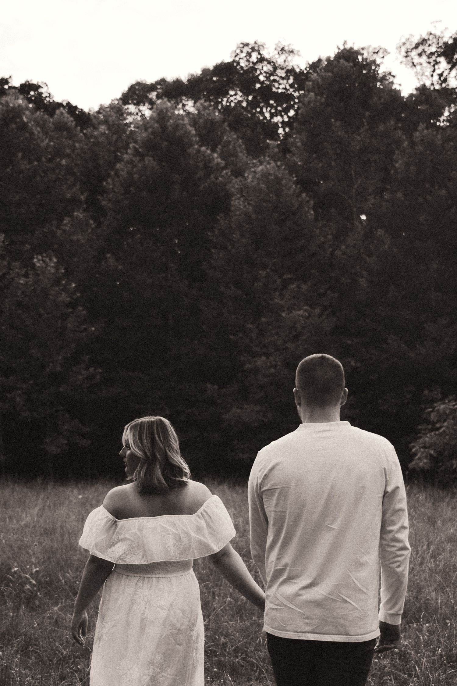Black and white photo of a couple standing in a grassy field at sunset holding hands against a backdrop of trees.