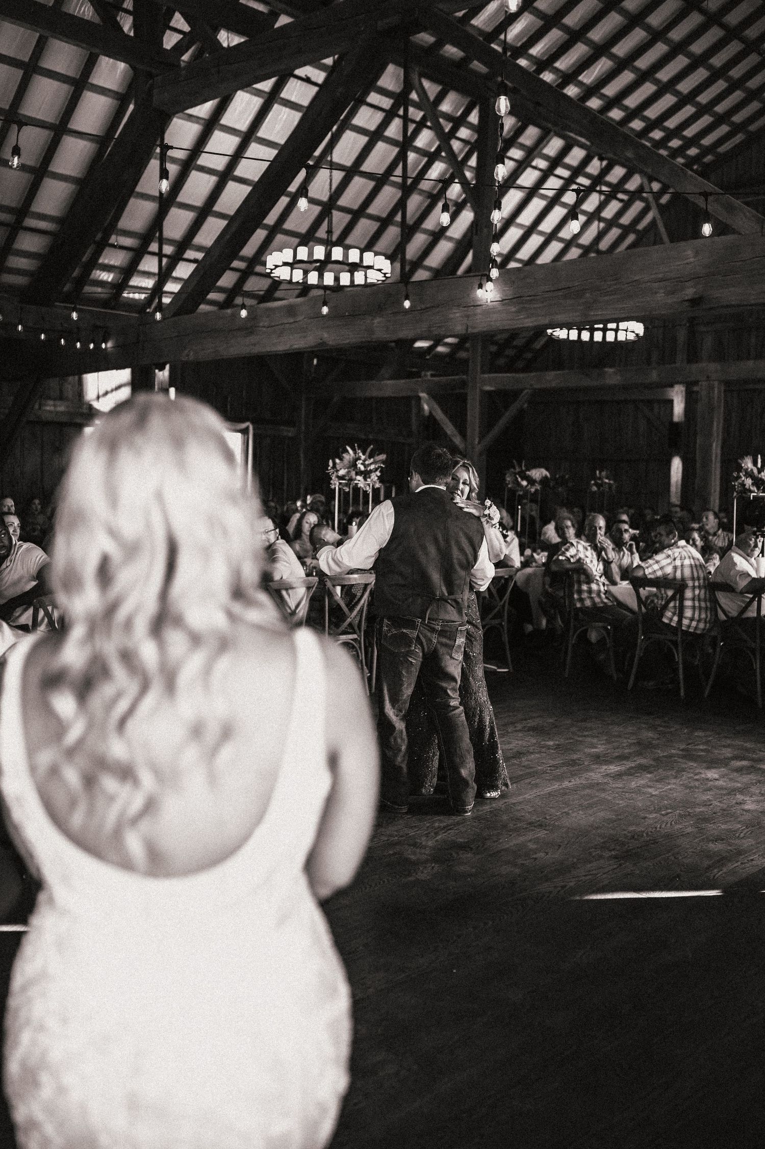 Black and white photo of a wedding reception in a rustic barn venue with exposed wooden beams and string lights.