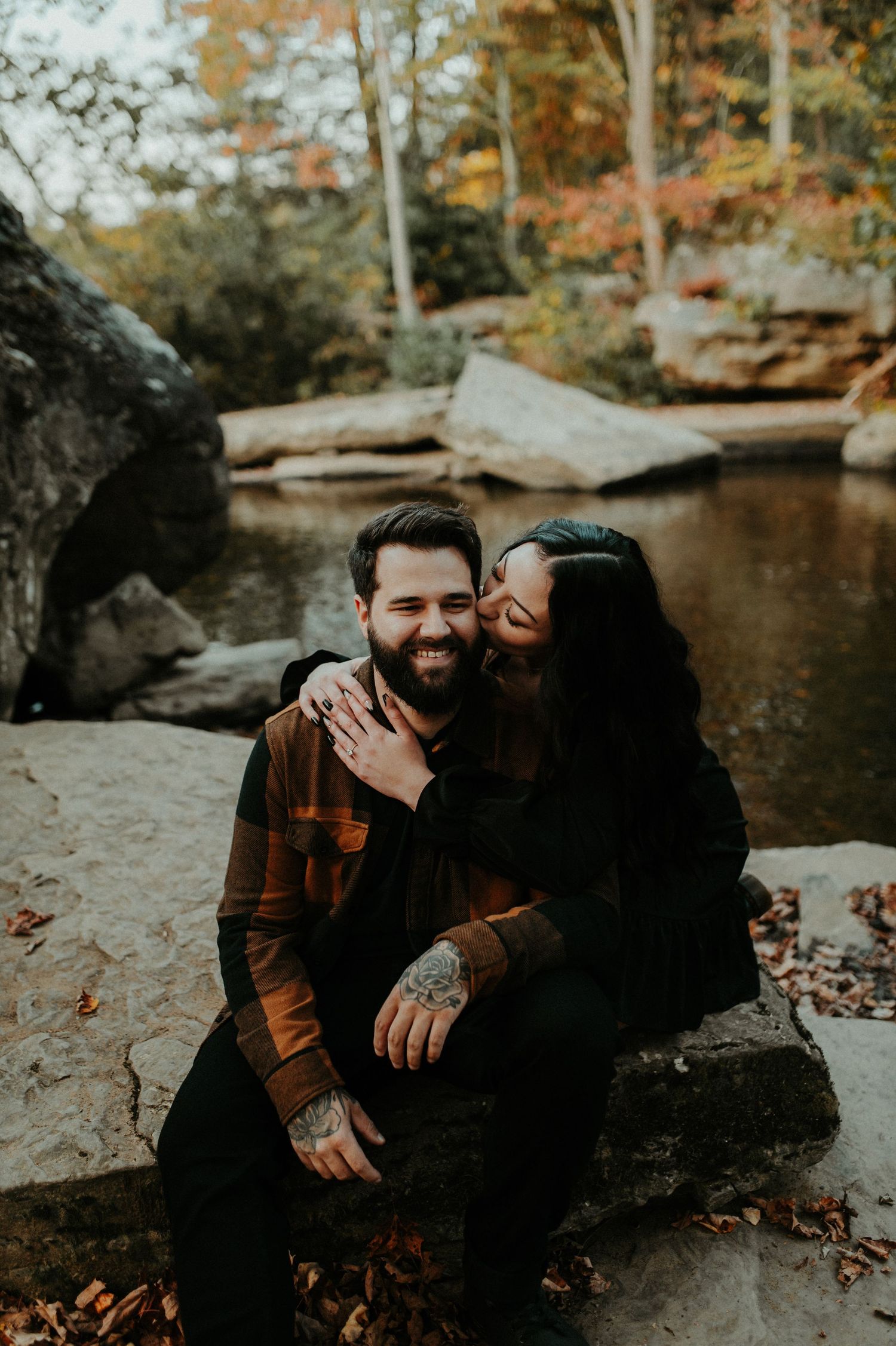Romantic autumn couple shares tender moment while sitting on rocks near a tranquil pond surrounded by fall foliage.