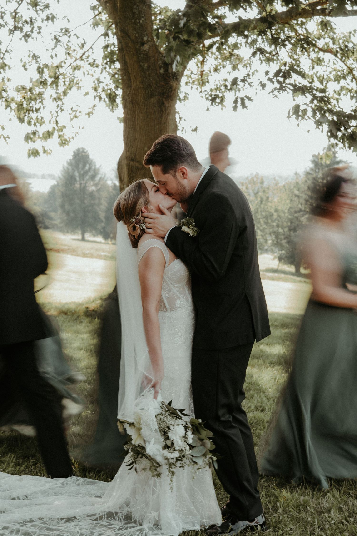 A romantic wedding moment under a tree with a couple in formal attire sharing a kiss while guests blur in motion around them.