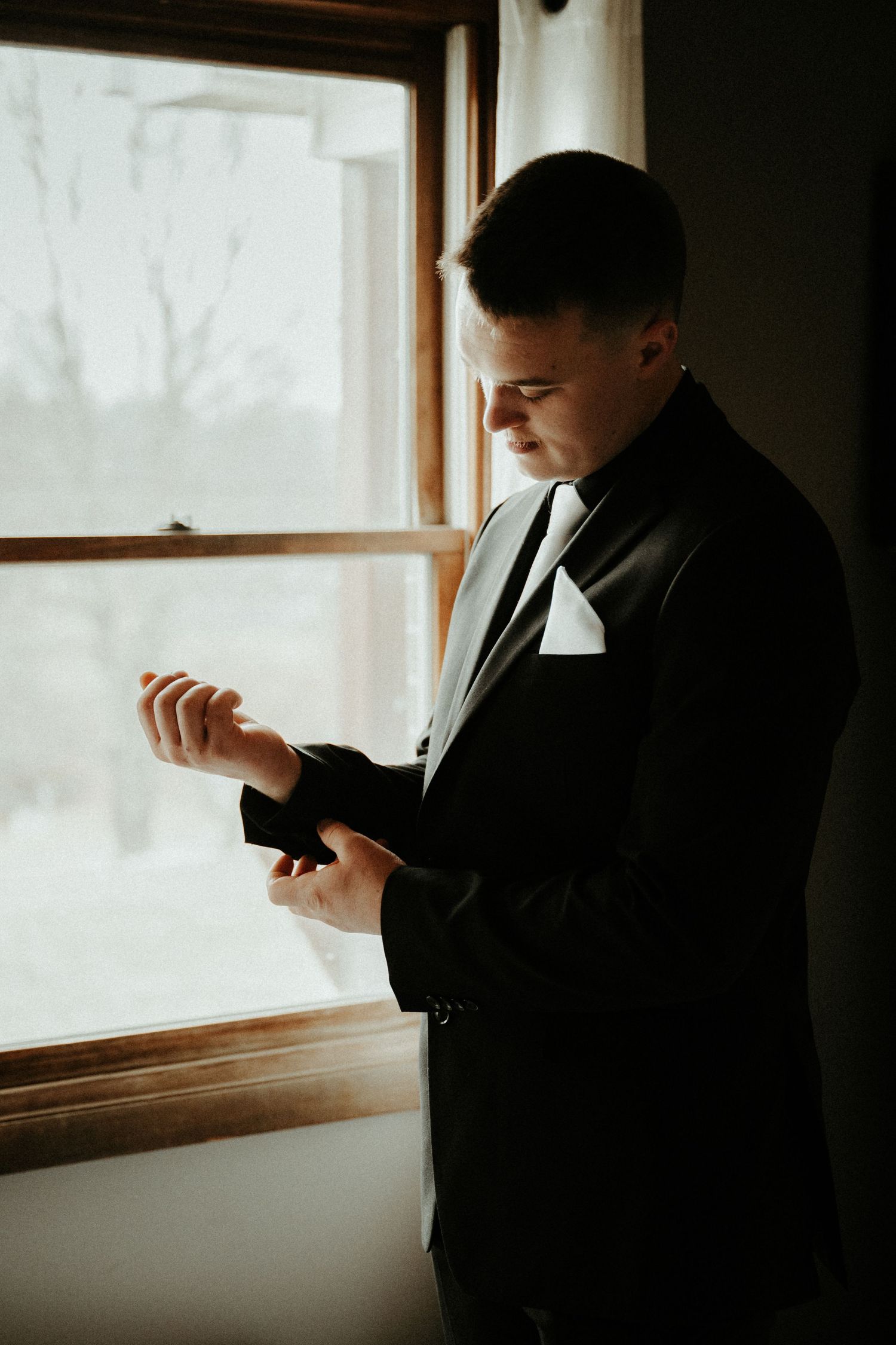 Person in formal black suit adjusts cufflinks while standing by a bright window. Pittsburgh Wedding Photographer
